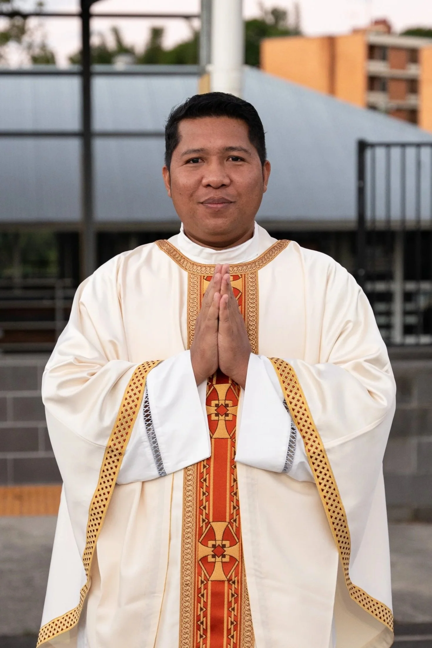A Catholic priest in a red religious robe speaking into a microphone at a podium, with a brick wall in the background.