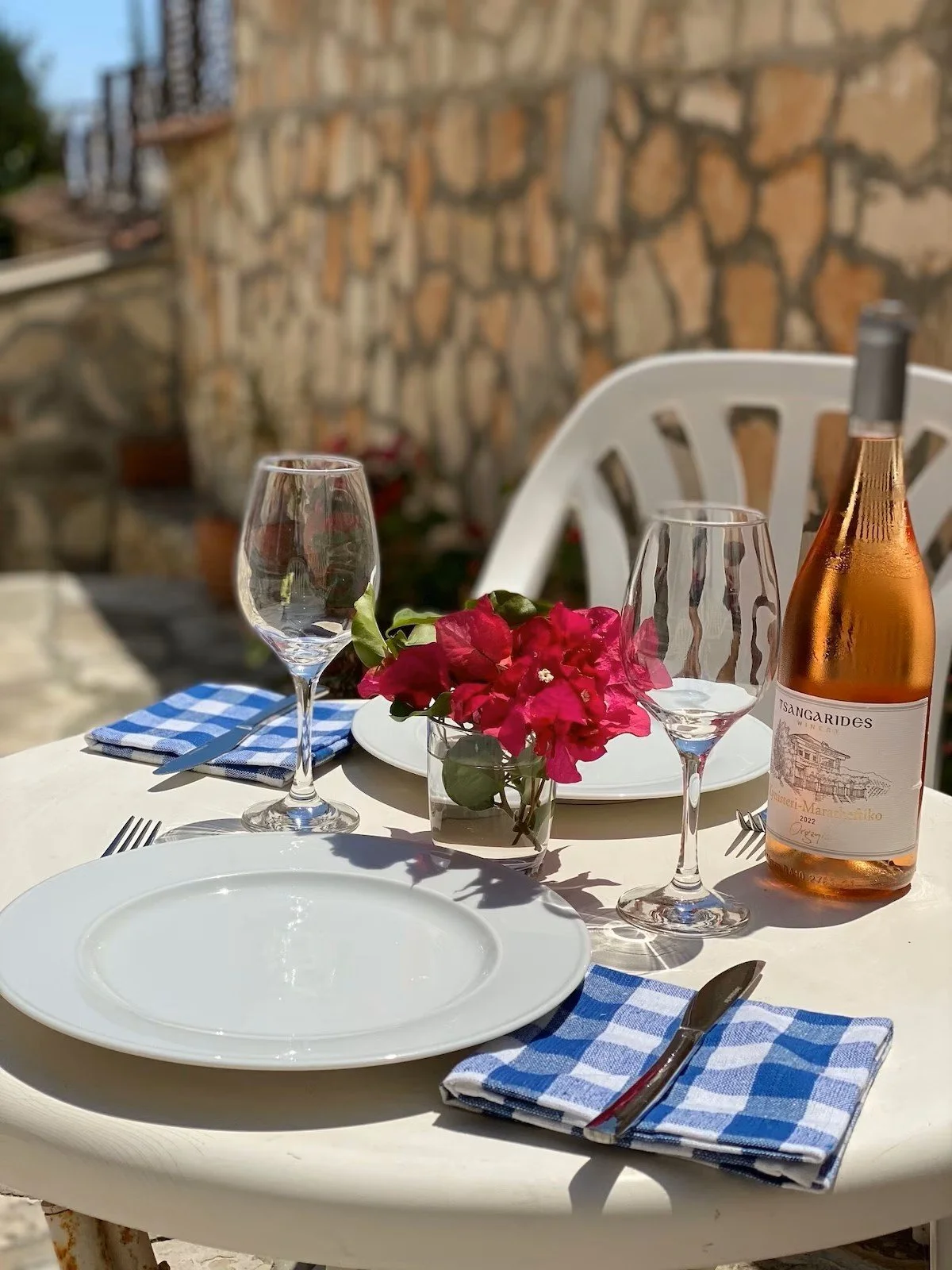 An outdoor dining table set with white plates, glasses, blue checkered cloth napkins with silverware, a small vase with pink flowers, and a bottle of rosé wine, against a stone wall background.