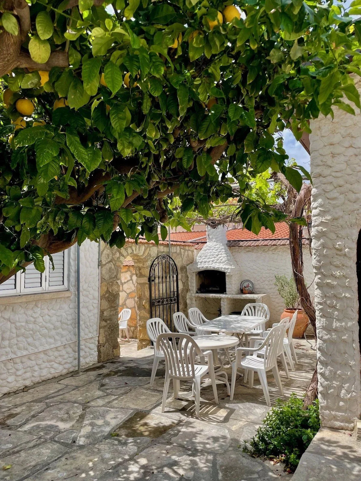 An outdoor patio area with a stone floor, surrounded by white textured walls and lush green trees. There is a white plastic dining table with six matching chairs. A stone arch and a black metal gate lead into another area. A white built-in outdoor fi