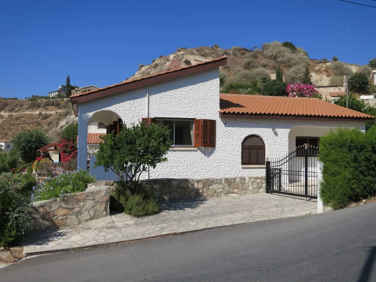 A white Mediterranean-style house with a red-tiled roof, surrounded by greenery and flowering plants, on a paved street with a rocky hillside in the background.