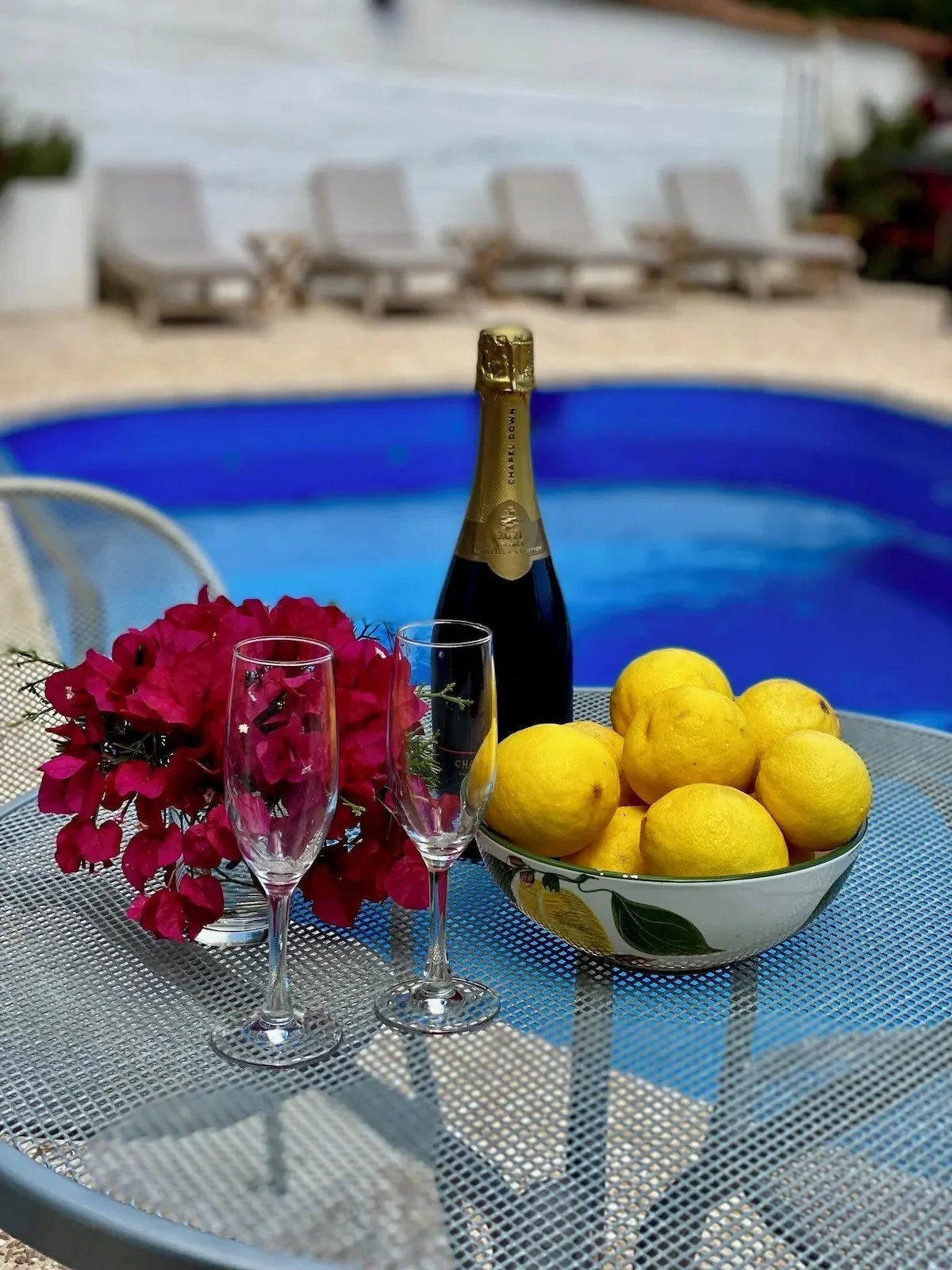 A table near a swimming pool with a bottle of champagne, two empty champagne glasses, a bowl of lemons, and a bouquet of pink flowers.