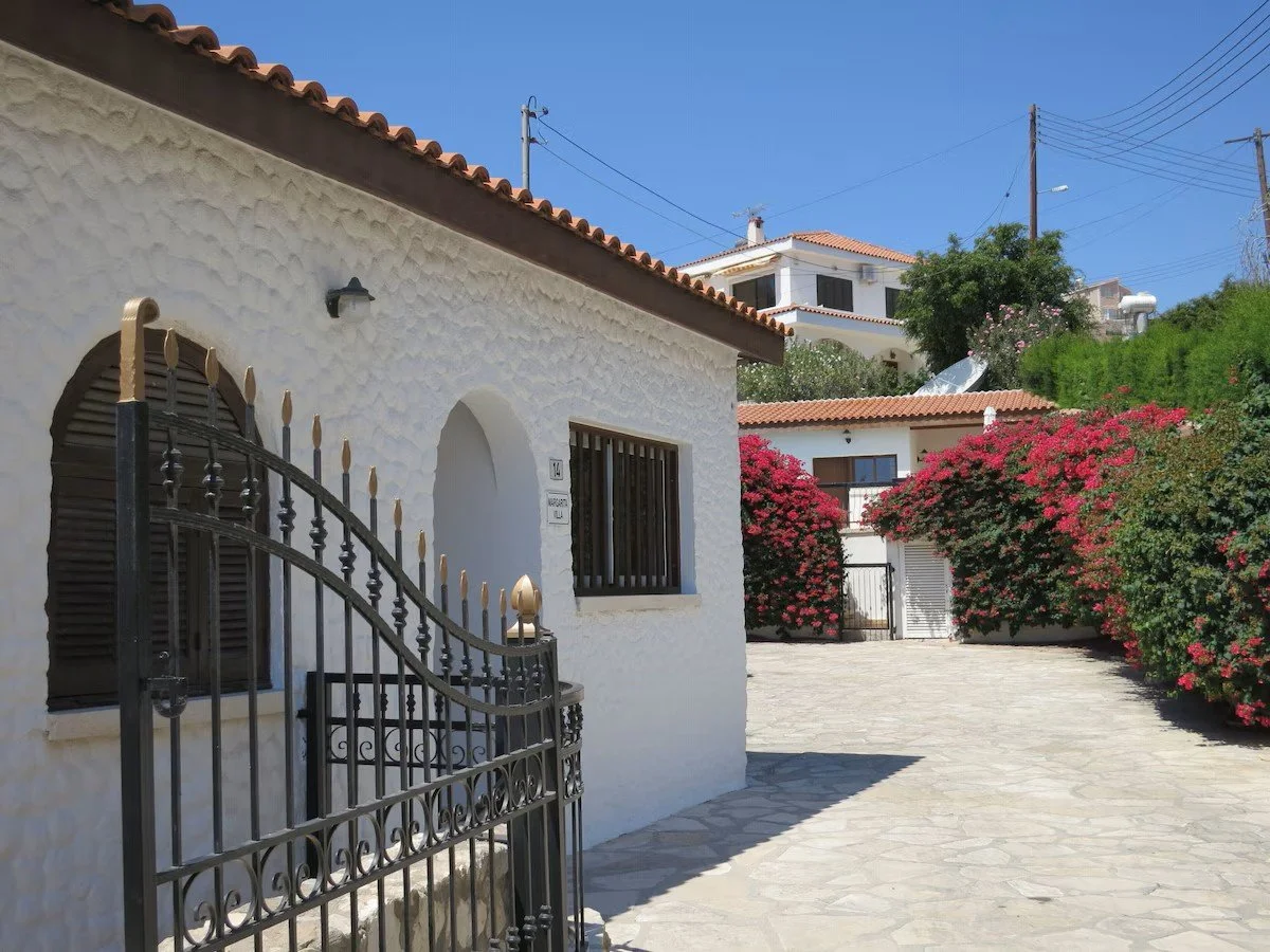 White villa with brown tiled roof, black iron gate, surrounded by pink flowering bushes