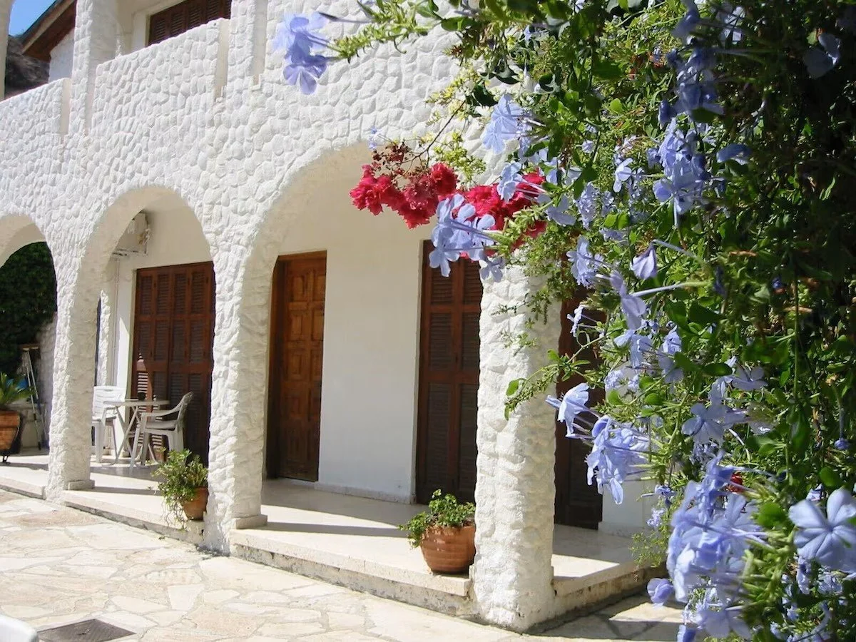 White building facade with stone arches, wooden shutters, and hanging potted plants, with colorful flowers in the foreground illustrating the lower terrace area