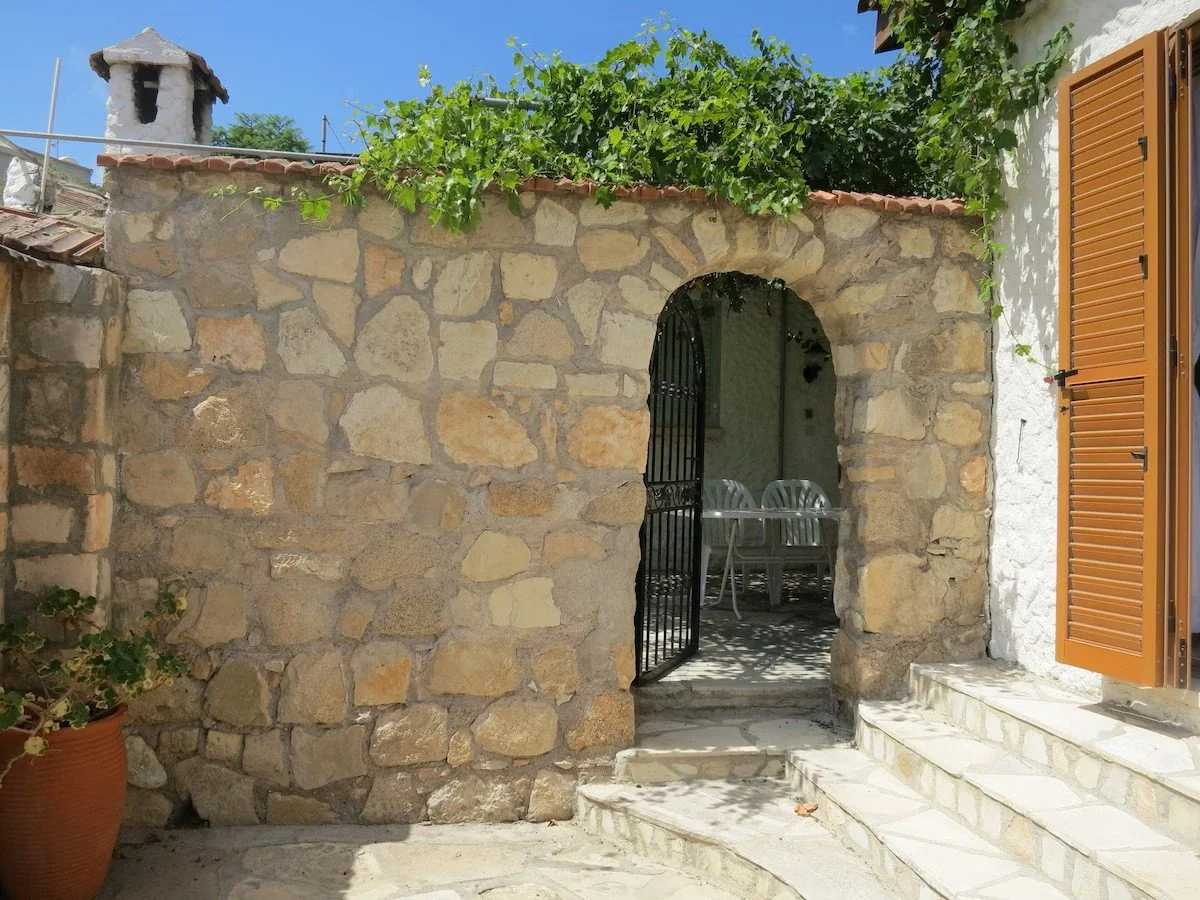 Stone archway with a black iron gate leading to a patio with white plastic chairs and table, surrounded by stone and white stucco walls, and green vines overhead.
