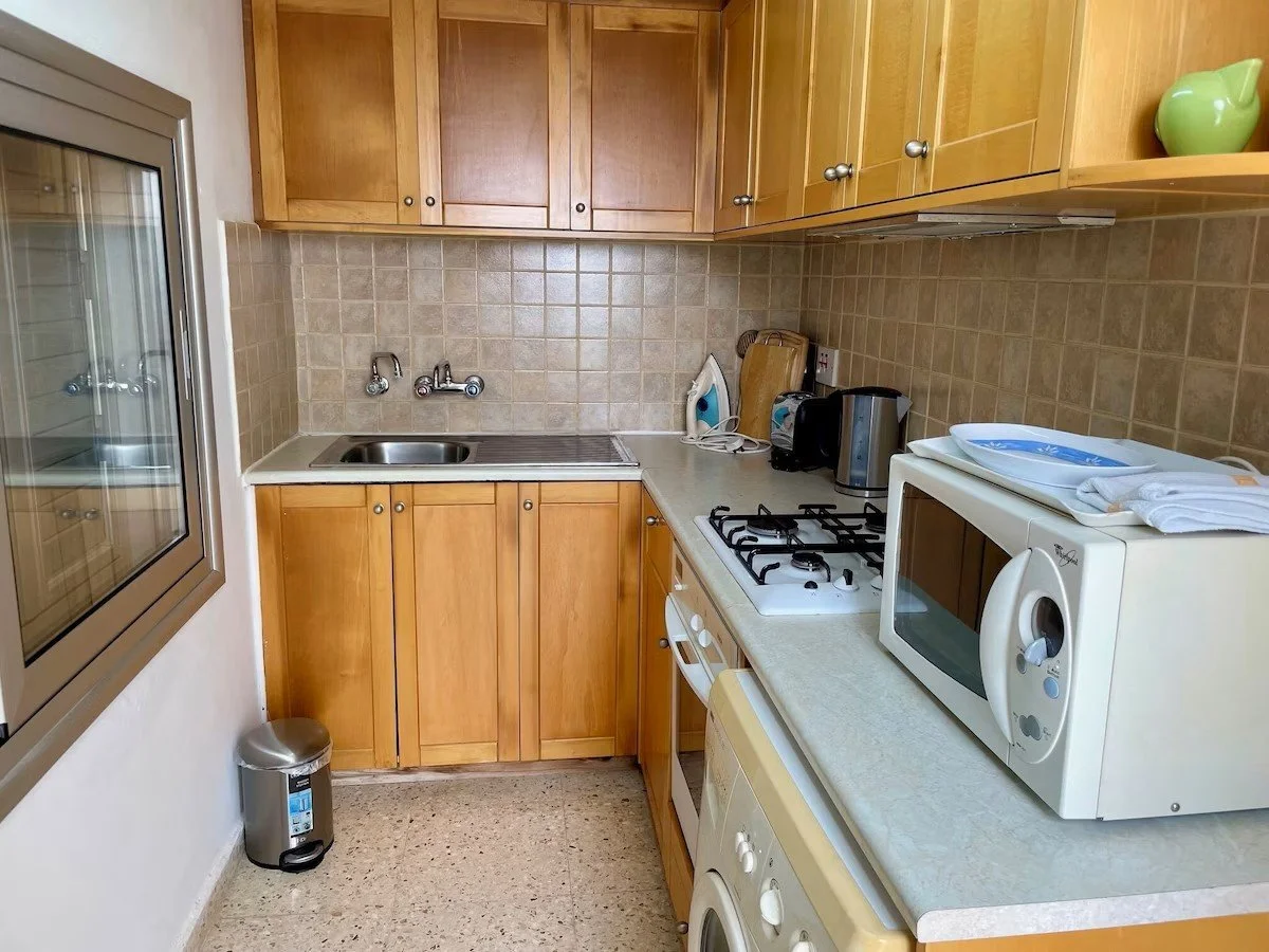 Compact kitchen with wooden cabinets, beige tile backsplash, stainless steel sink, electric stove, microwave, toaster, iron, and a window on the left.