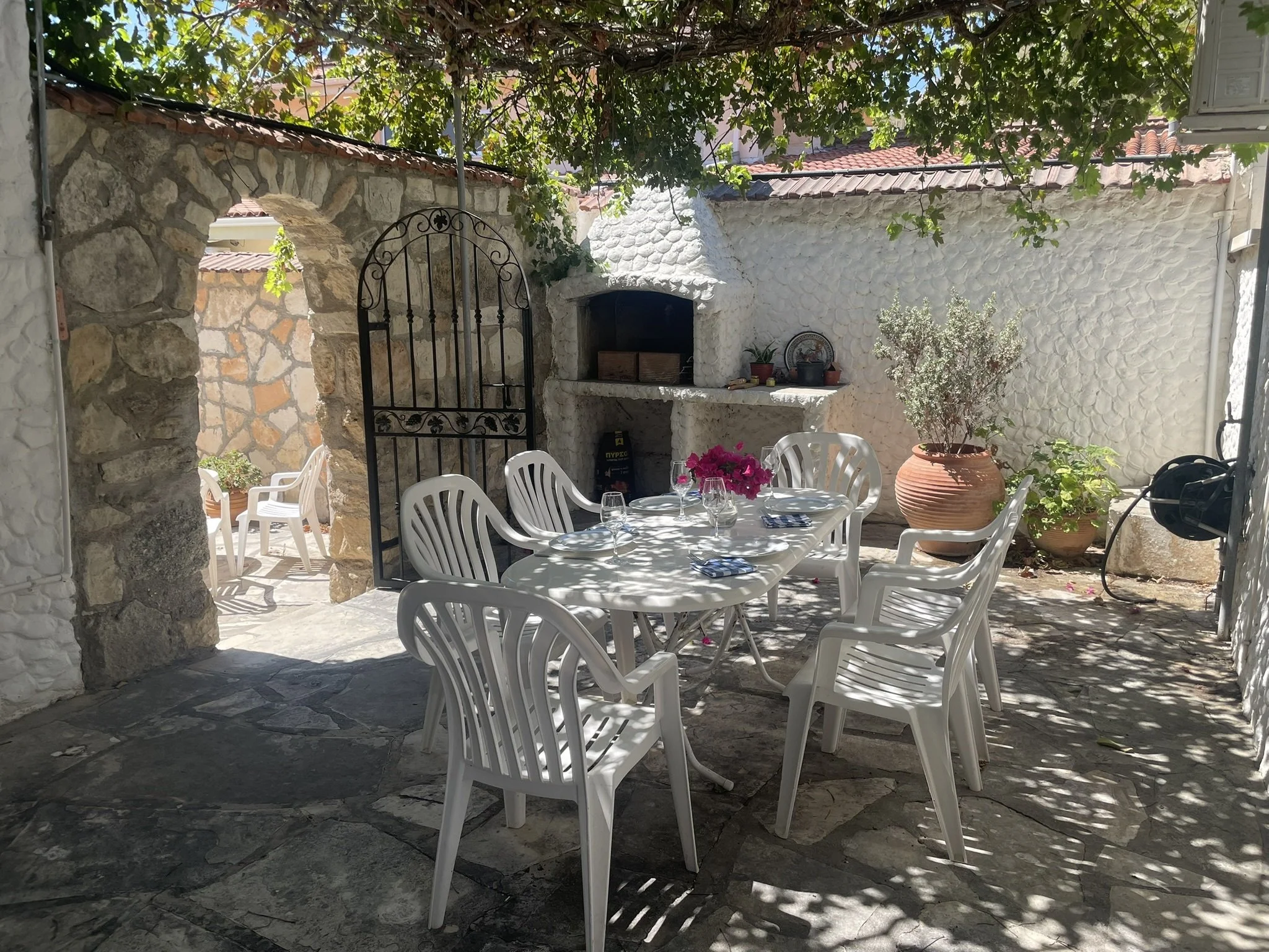 An outdoor patio with a white table and eight chairs, under dappled shade with lunch set for four people