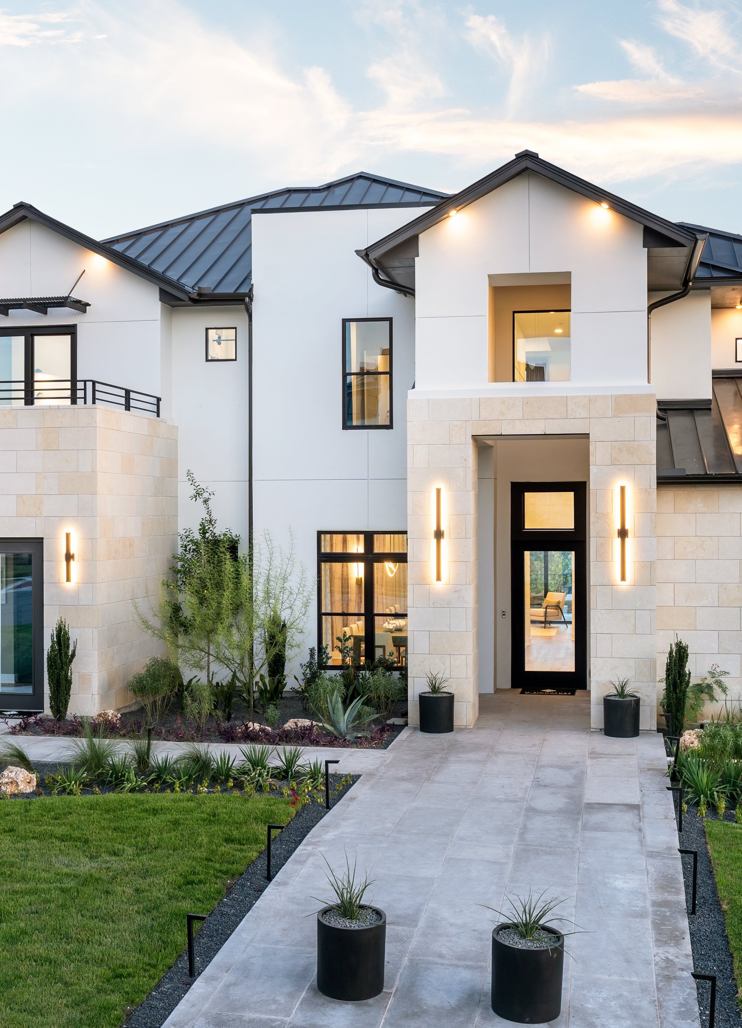 Modern house with a landscaped front yard, large windows, a black door, exterior lighting, and potted plants along a stone walkway under a partly cloudy sky in the evening.