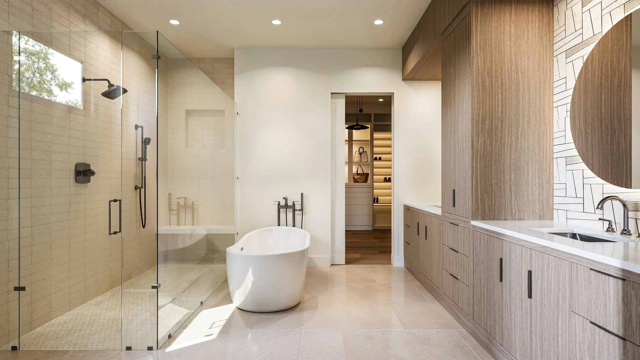 Modern bathroom with glass-enclosed shower, freestanding bathtub, wooden vanity with double sink, and walk-in closet visible through an open door.