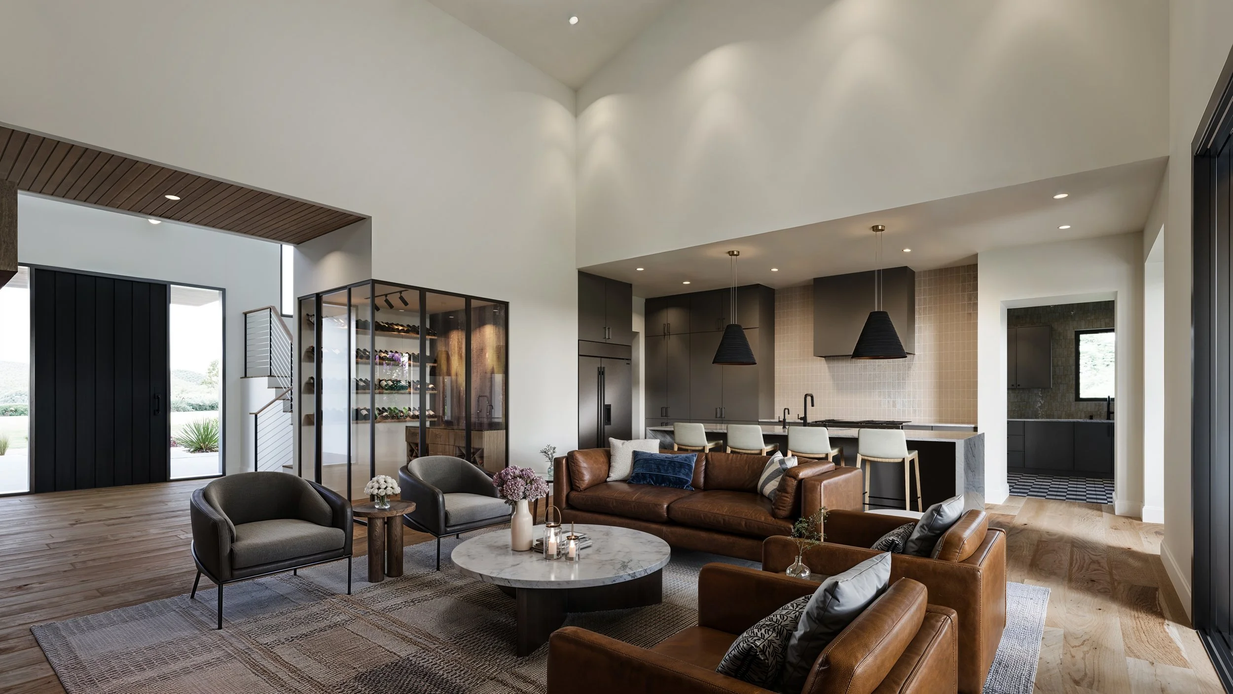 Modern open-concept living room and kitchen with high vaulted ceiling, neutral tones, pendant lights, leather sofas, and a wine display.
