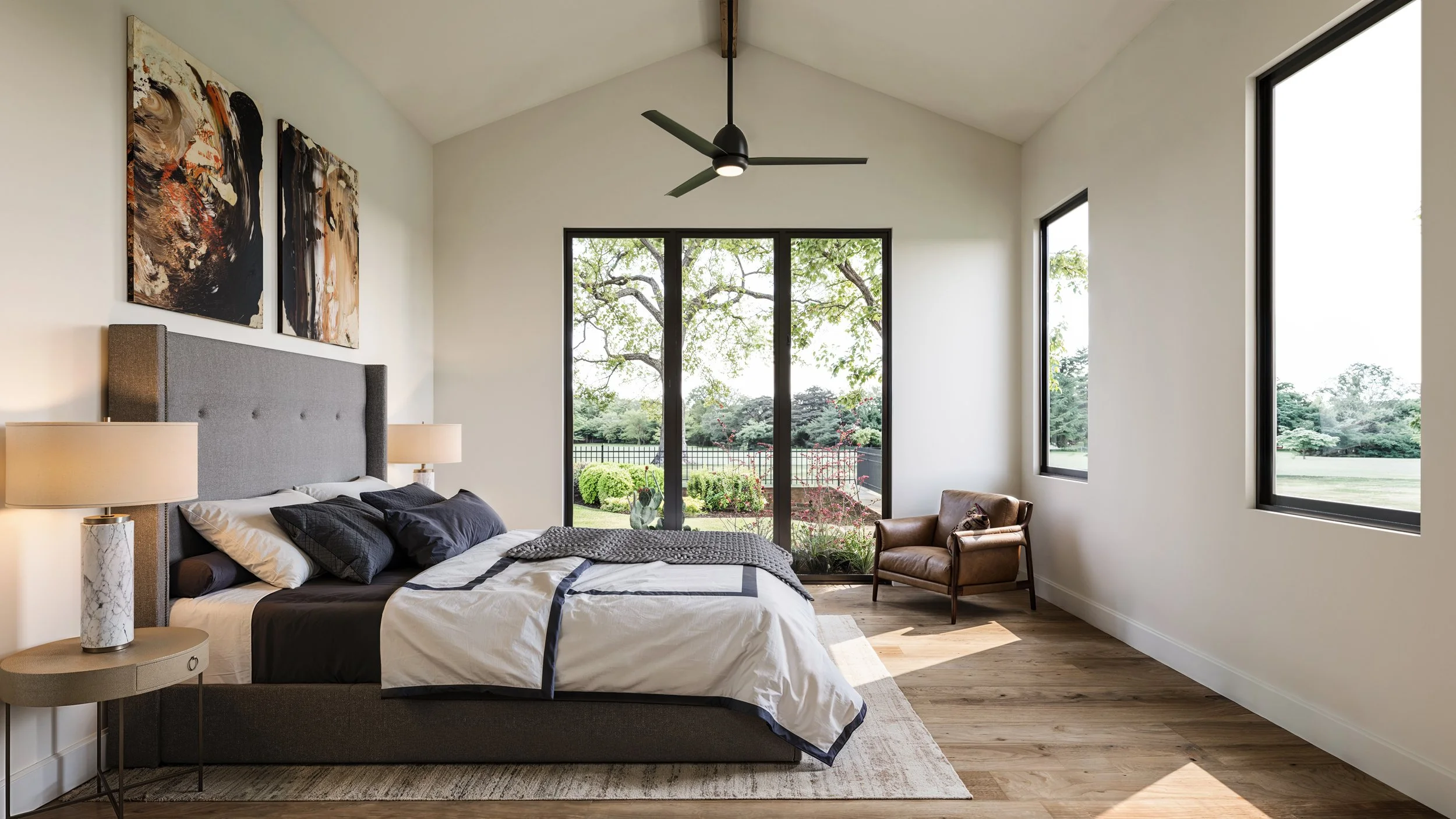 Modern bedroom with a large bed, black, white, and gray bedding, and a gray headboard. Two bedside lamps with marble bases are on either side of the bed. Large windows and a glass door show a lush outdoor landscape with trees and bushes. Abstract art