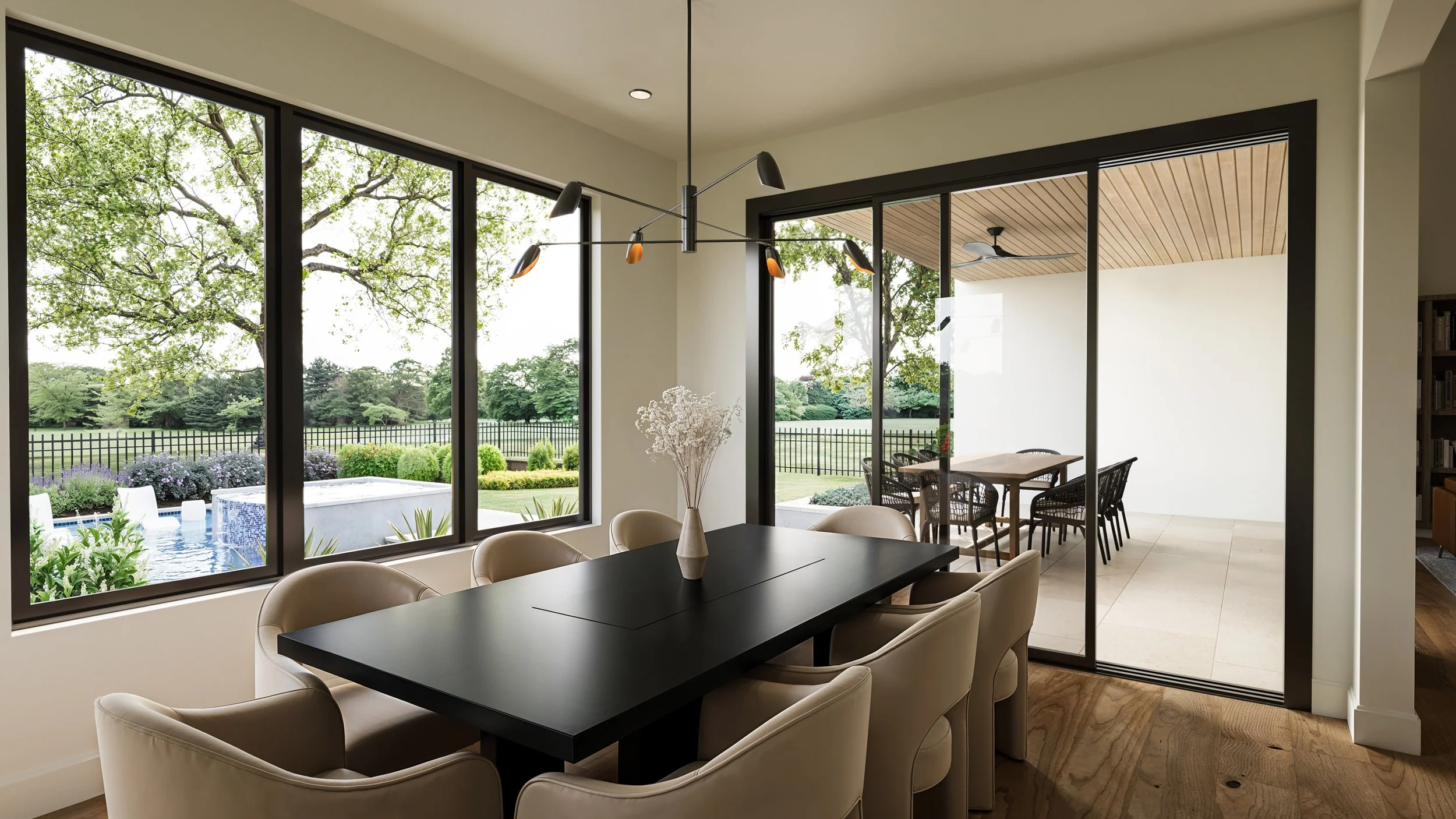Interior of a modern dining room with large windows showcasing a garden and swimming pool outside. The room features a black dining table surrounded by cream-colored chairs, a minimalist chandelier, and a small balcony or patio area with outdoor furn