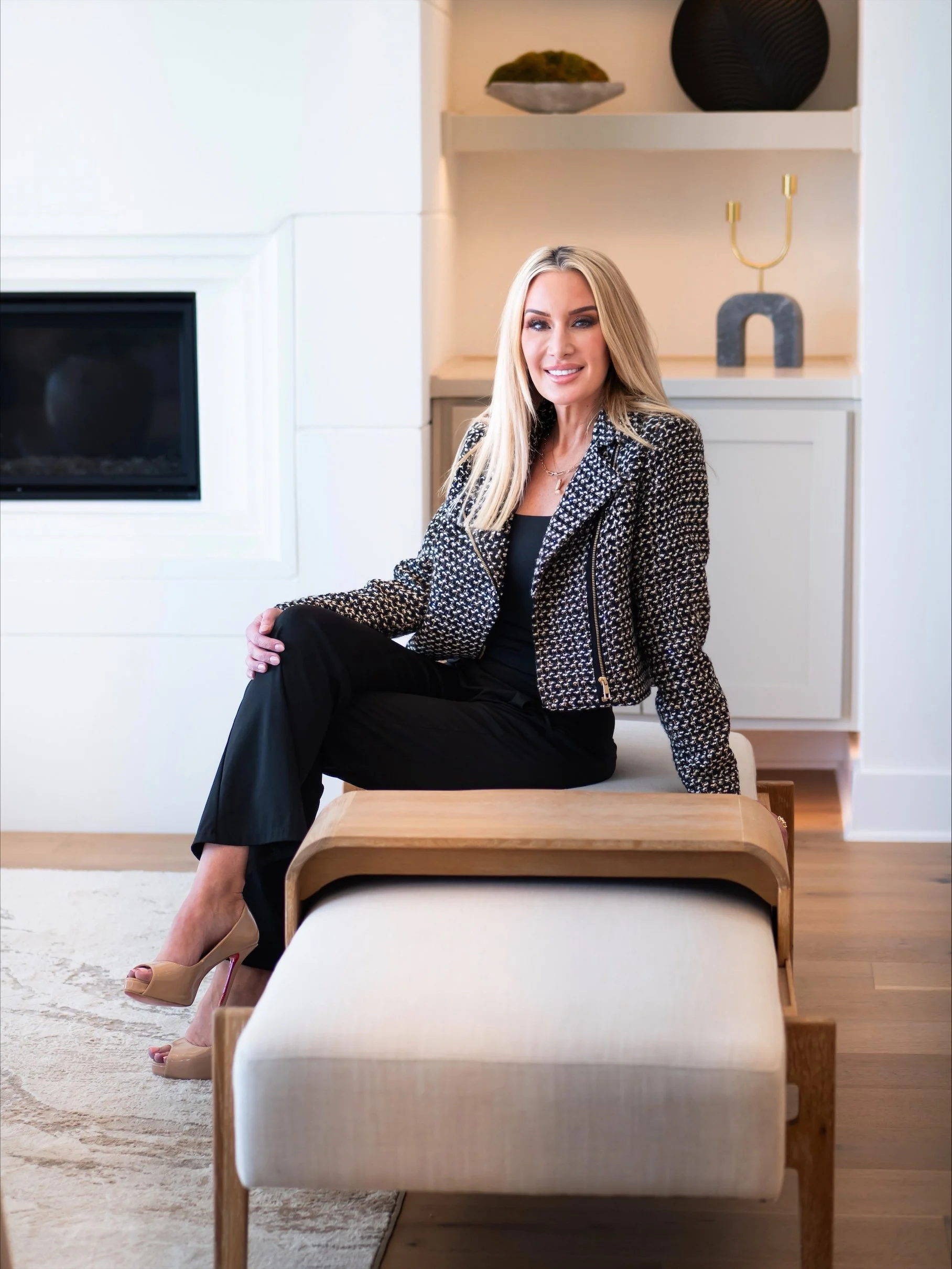 A woman with long blonde hair sitting on a beige ottoman in a modern living room, wearing a black dress, a patterned jacket, and beige high heels, smiling at the camera.