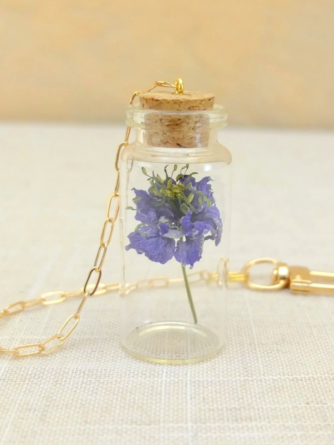 Glass bottle with a cork lid containing a purple flower, gold chain accessory, on a light fabric surface. A fable and twig hanging tiny jar