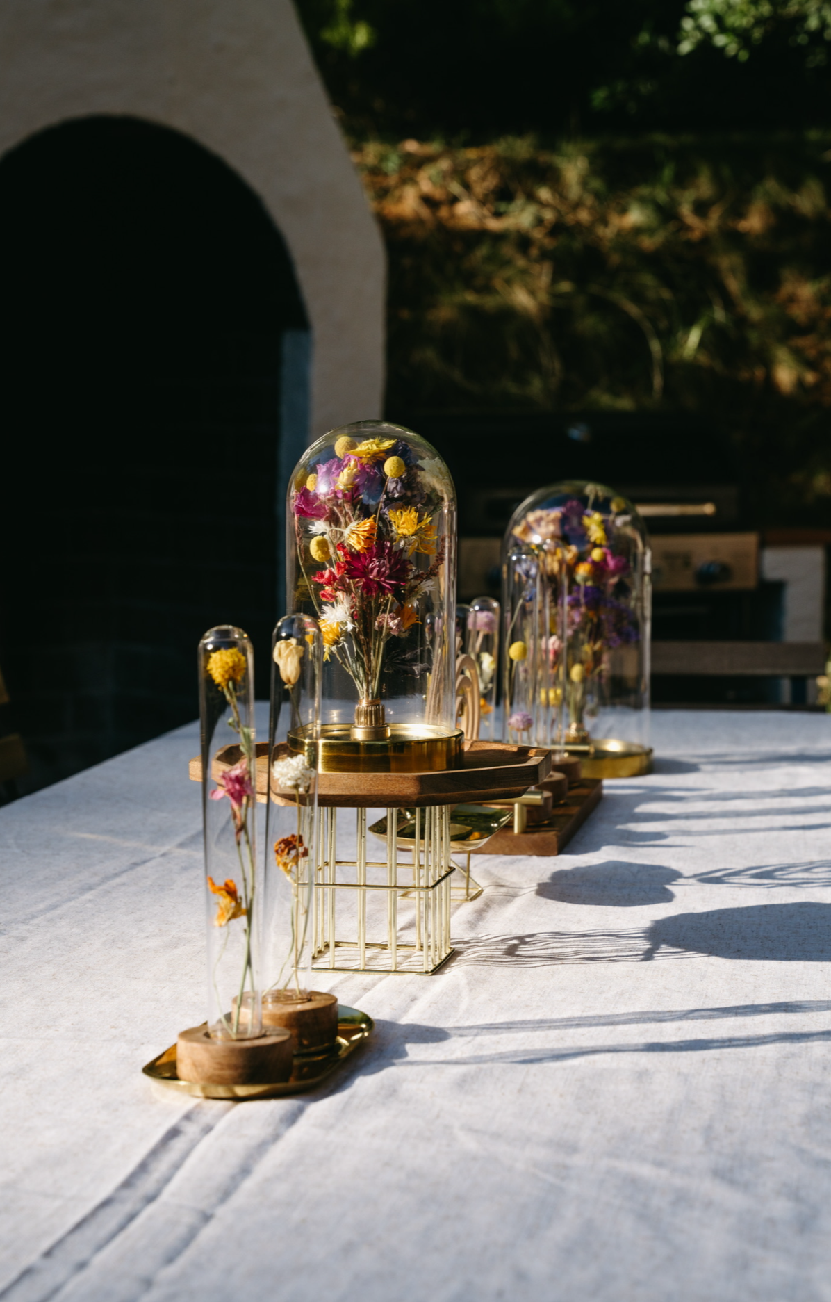 Elegant outdoor table setting with glass display domes containing colorful dried flowers, arranged on wooden and metal stands, casting long shadows on a white tablecloth. An elopement or micro wedding styled by fable and twig’s hire service.