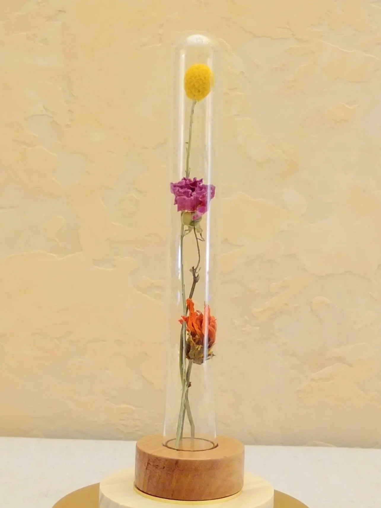 A glass tube with dried flowers in pink, orange, and yellow, mounted on a wooden base. A fable and twig test tube with custom timber and brass base