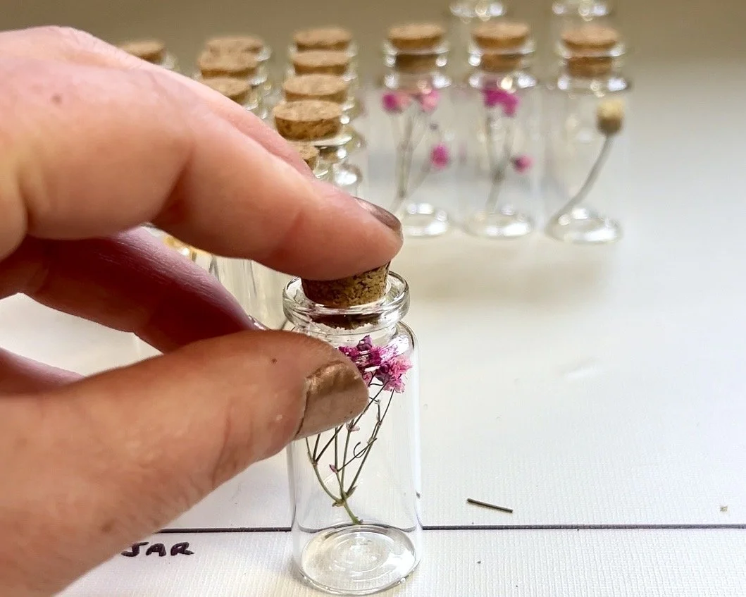 A hand placing a cork into a small glass jar with pink babies breath flowers inside, with other similar tiny vials lined up in the background.