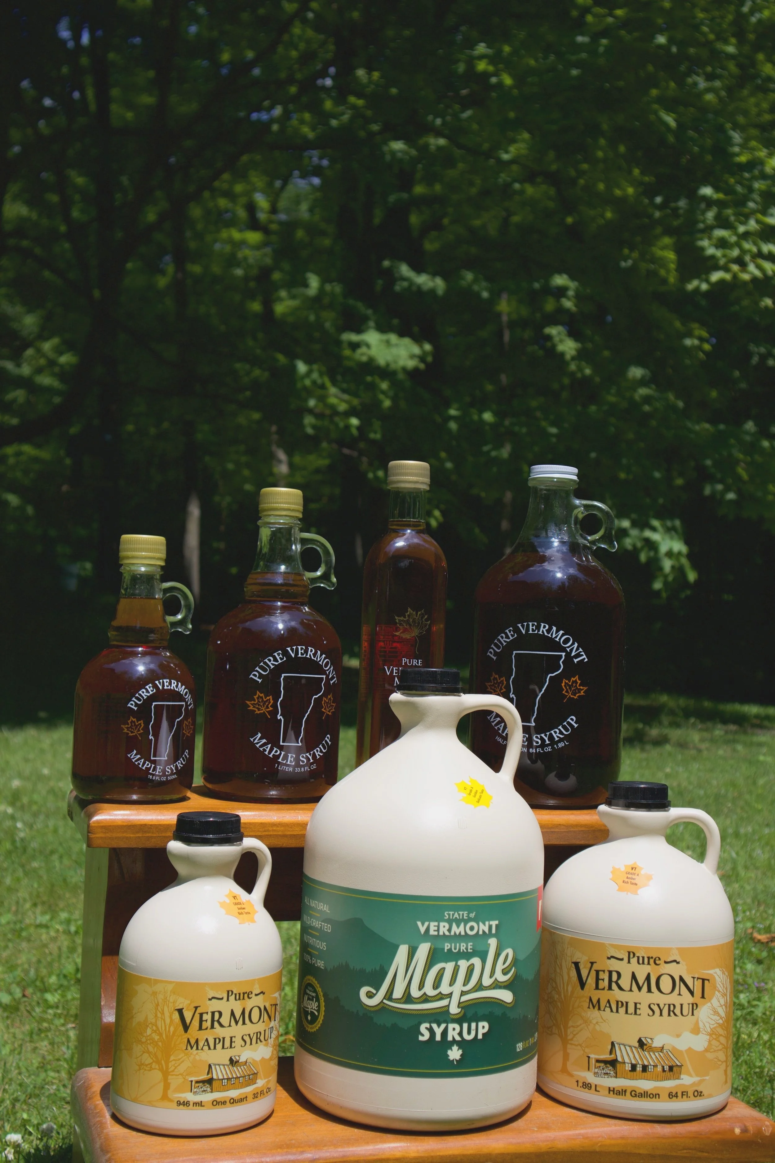 Collection of Vermont maple syrup bottles on a wooden stand outdoors with trees in the background.