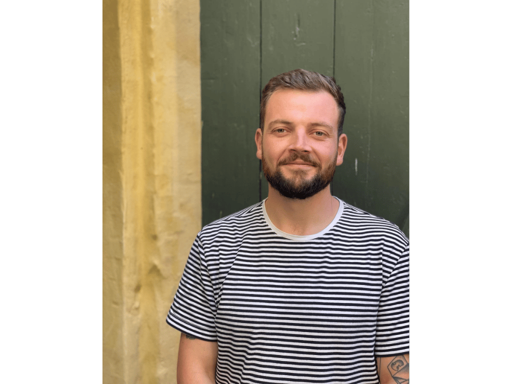 A man with short hair and a beard, wearing a striped t-shirt, smiling outdoors in front of a green and yellow wall.