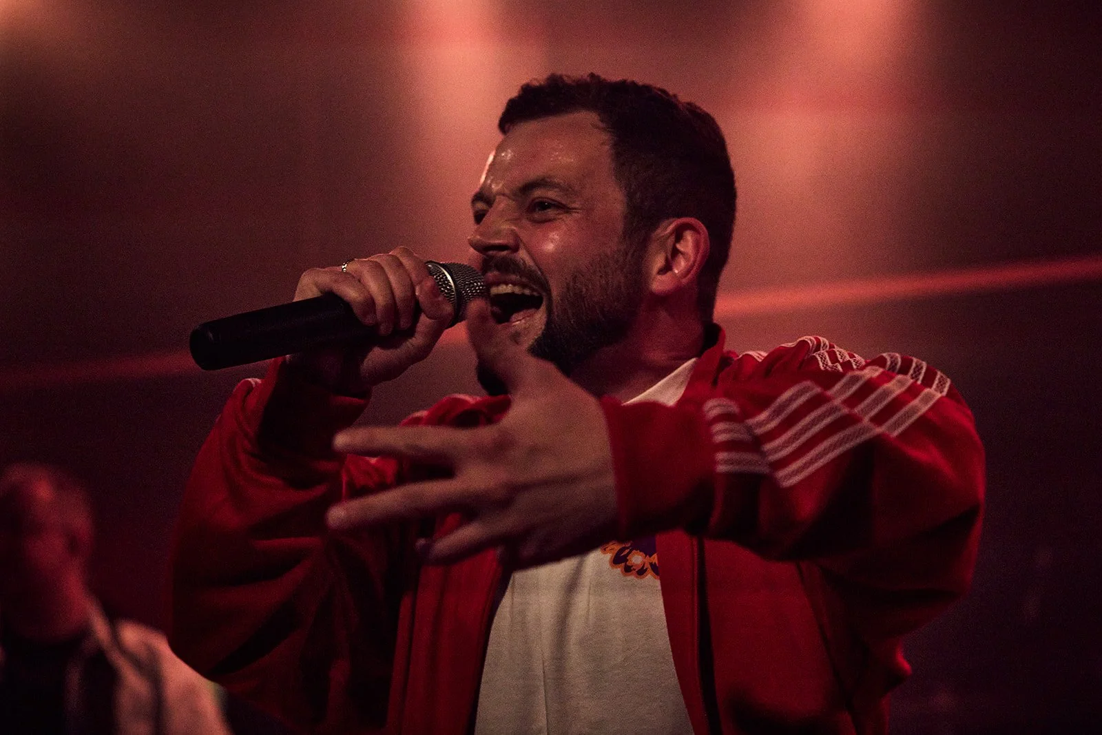 A man singing into a microphone with a big smile, wearing a red jacket with white details, in a dimly lit setting. Ideal Bar, Vega, Frit Løb, Cillian Mxrphy