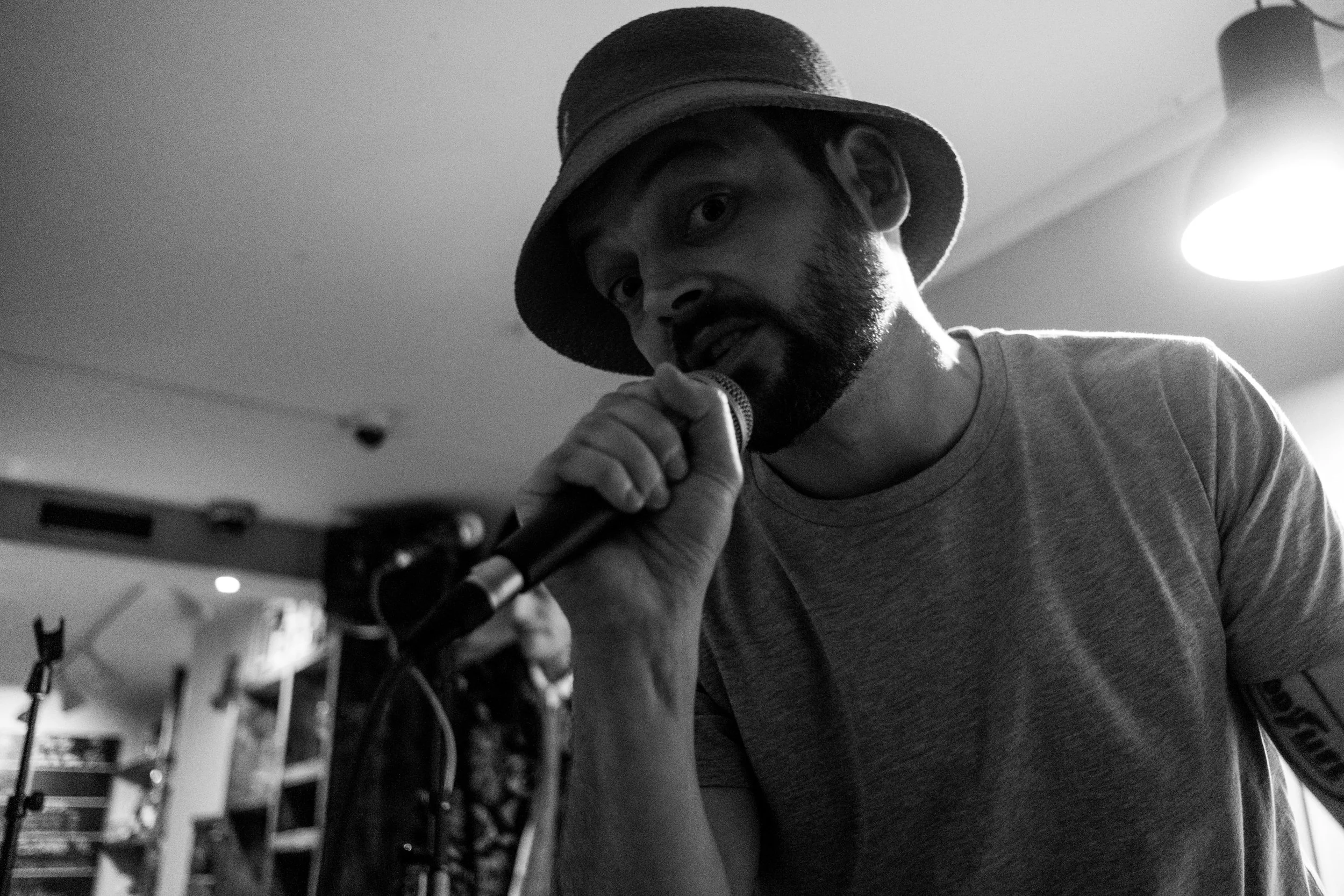 Man wearing a bucket hat holding a microphone close to his mouth, looking into the camera in black and white, indoors with shelves and equipment in the background. Billede af Tinus Buus.