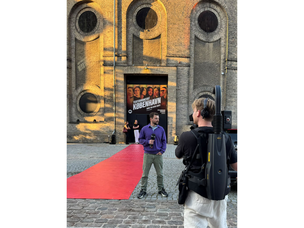 A man being interviewed on a red carpet outside a theater with a large poster for a concert called 'København.' A cameraman with a shoulder-mounted camera films the interview. Østre Gasværk, Gaffa.