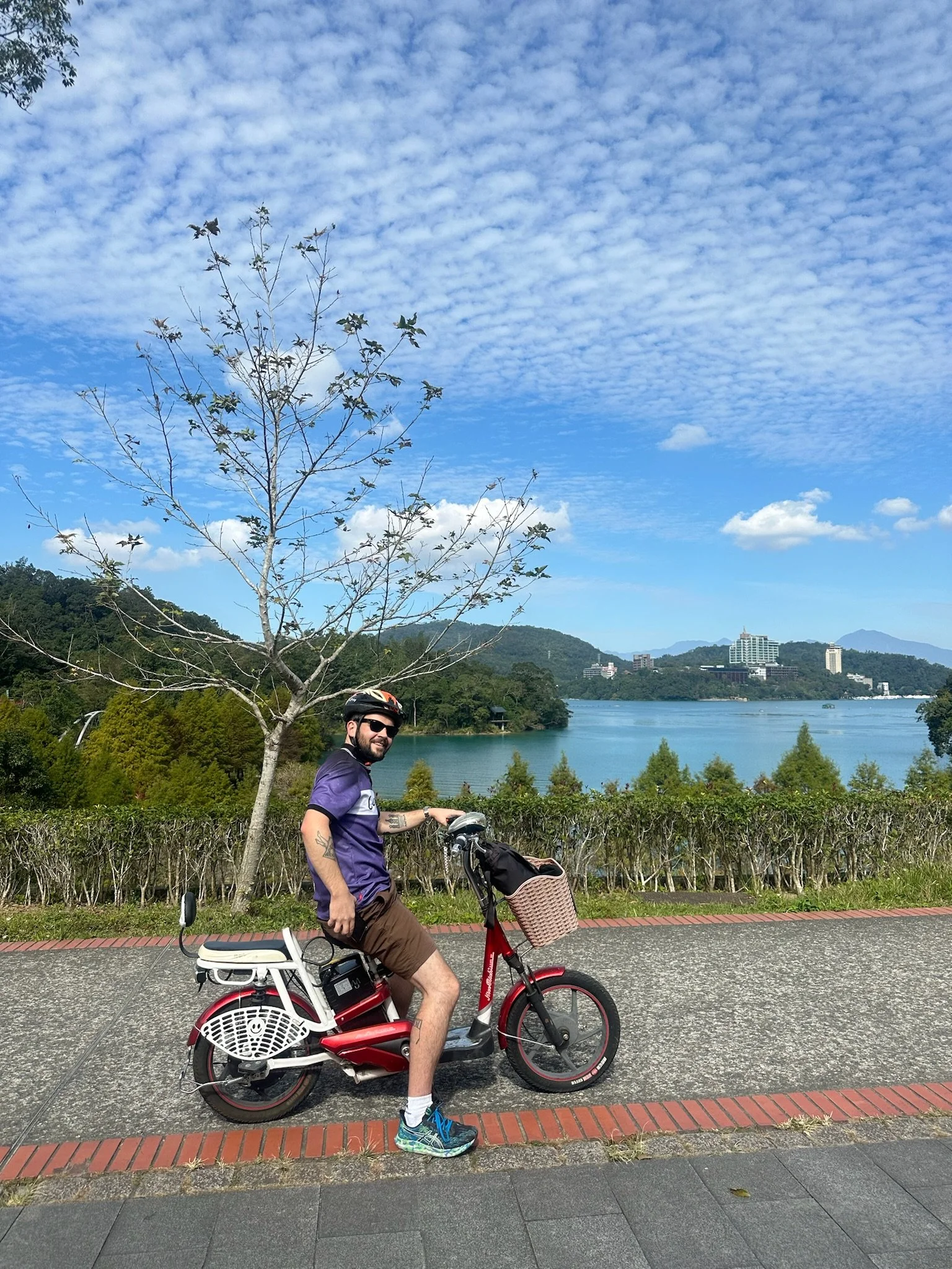 A man in a purple shirt and brown shorts riding a red electric bike along Sun Moon Lake, Taiwan with a tree, green bushes, and buildings in the background under a partly cloudy blue sky.