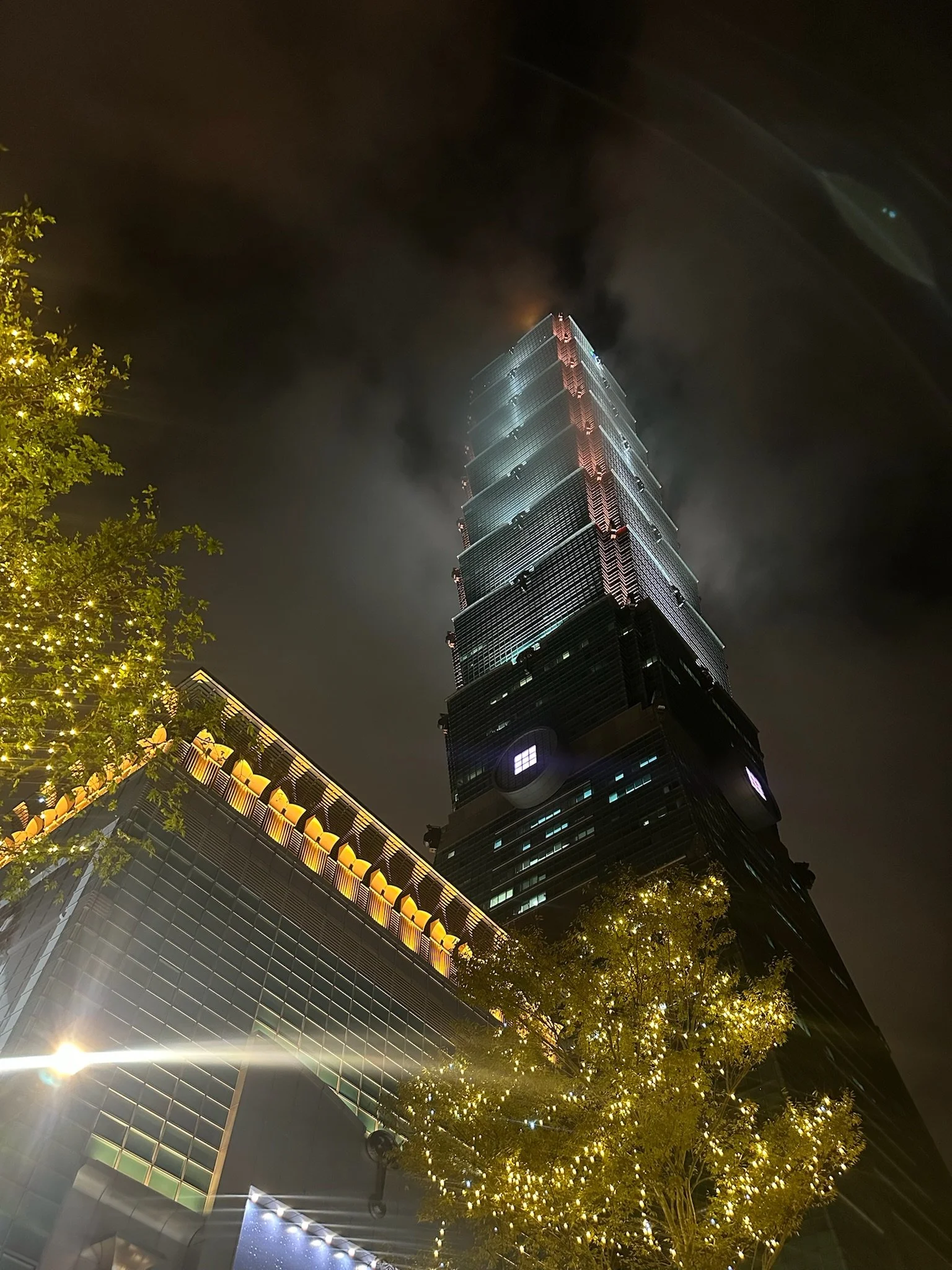 Night view of Taipei 101 skyscraper with illuminated exterior, surrounded by trees with festive string lights.
