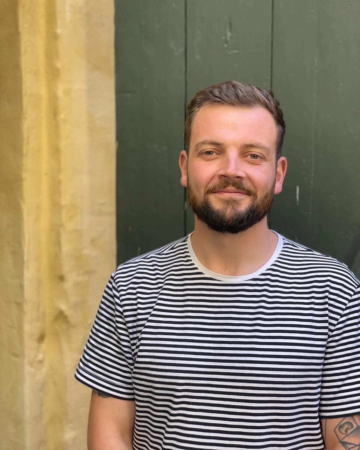 A young man with a beard and short brown hair is standing in front of a dark green door, smiling softly, wearing a black and white t-shirt.
En ung mand med skæg og kort brunt hår står foran en mørkegrøn dør med et mildt smil i en sort-hvid t-shirt