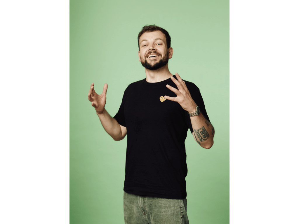 A man with a beard and short hair wearing a black T-shirt with a small embroidered logo on the chest, standing against a green background. He is smiling and gesturing with both hands. Rapolitics, Line Westerboe