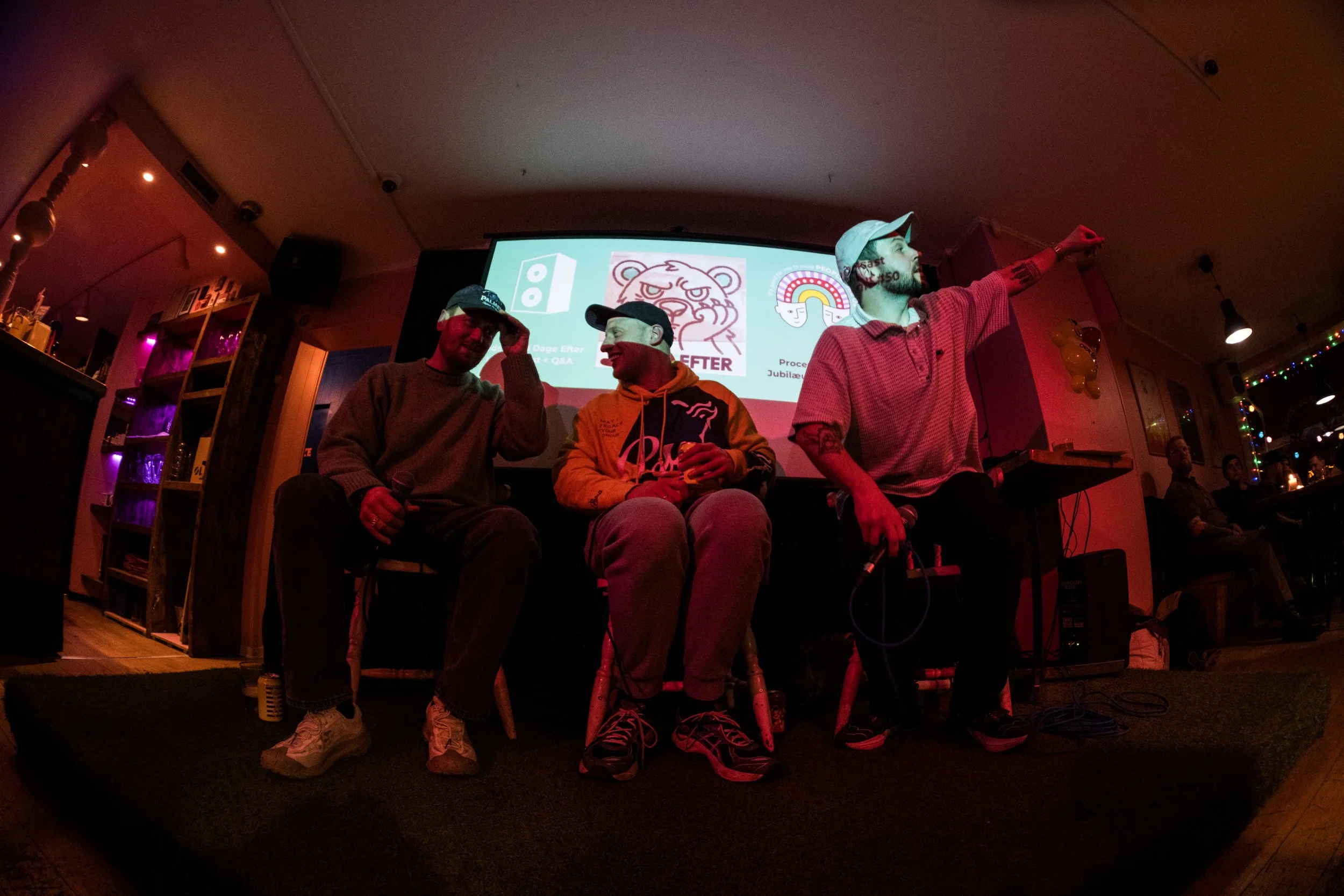 Three men sitting on a stage in a dimly lit room, with a woman in the background. The man on the left is wearing a gray sweater and a black cap, holding a microphone. The woman in the middle is smiling, wearing a hoodie and sneakers. The man on the r