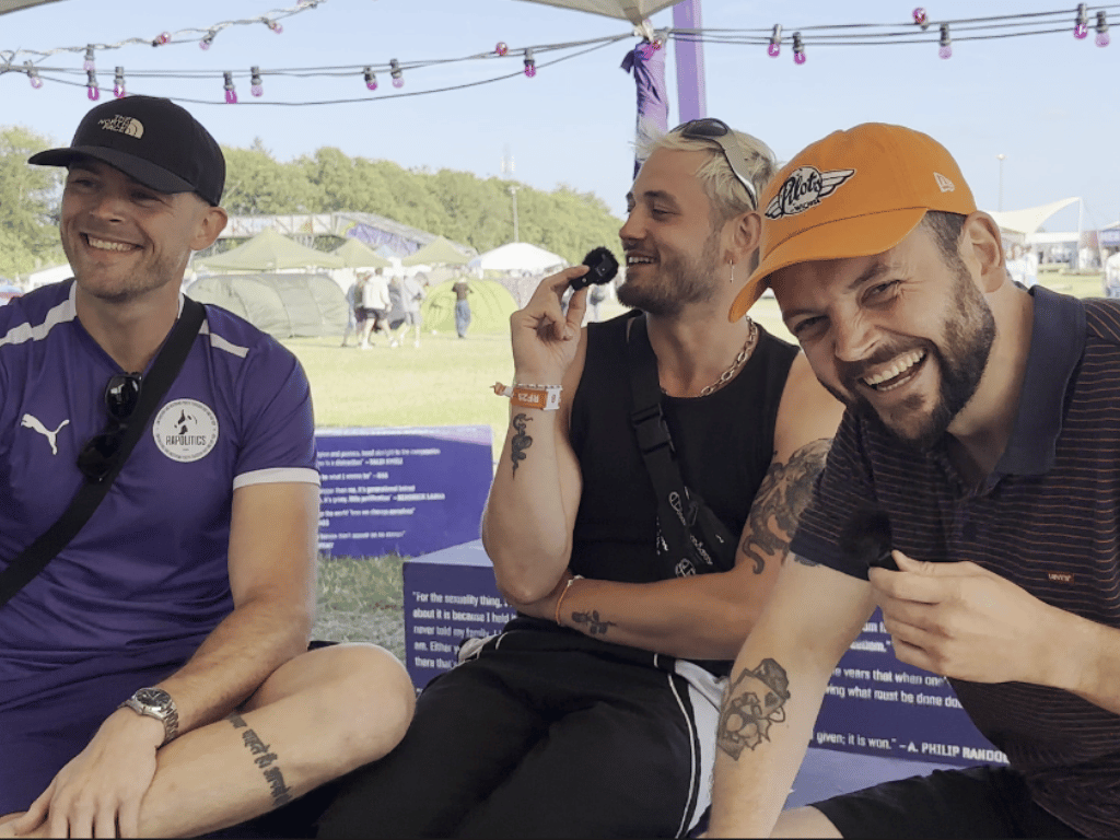 Three men sitting outdoors at a festival or outdoor event, smiling and laughing. They are wearing casual clothing, including hats and sunglasses. Tents and people in the background. Rapolitics, Re:Act, Roskilde Festival, Processen Bag..., Manus Bell