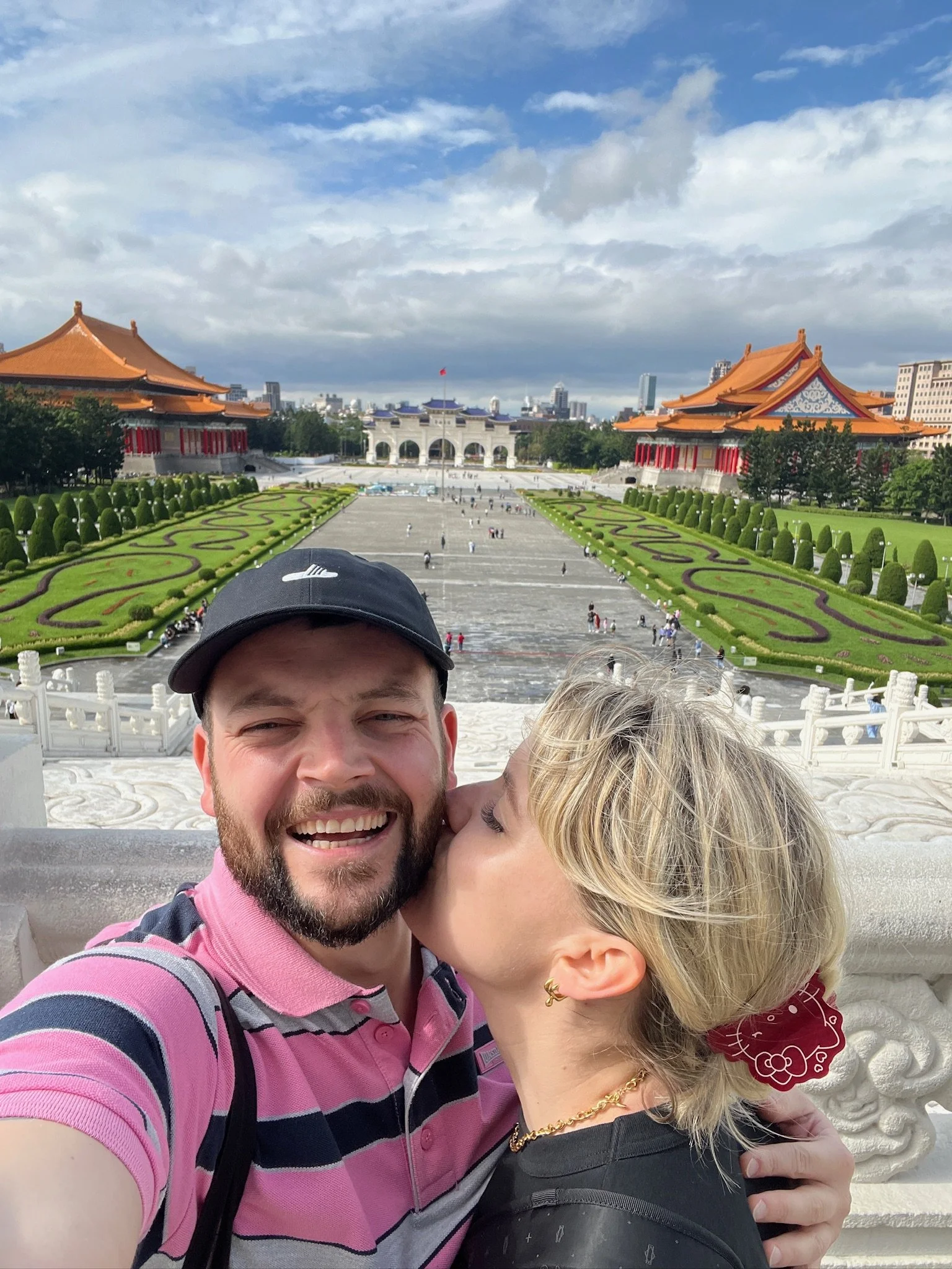 A smiling man in a pink striped polo shirt and a woman with blonde hair, gold jewelry, and a red Hello Kitty hair clip, sharing a kiss in front of the National Revolutionary Martyrs' Shrine with traditional Chinese architecture, green landscaped gardens, and a cloudy blue sky.