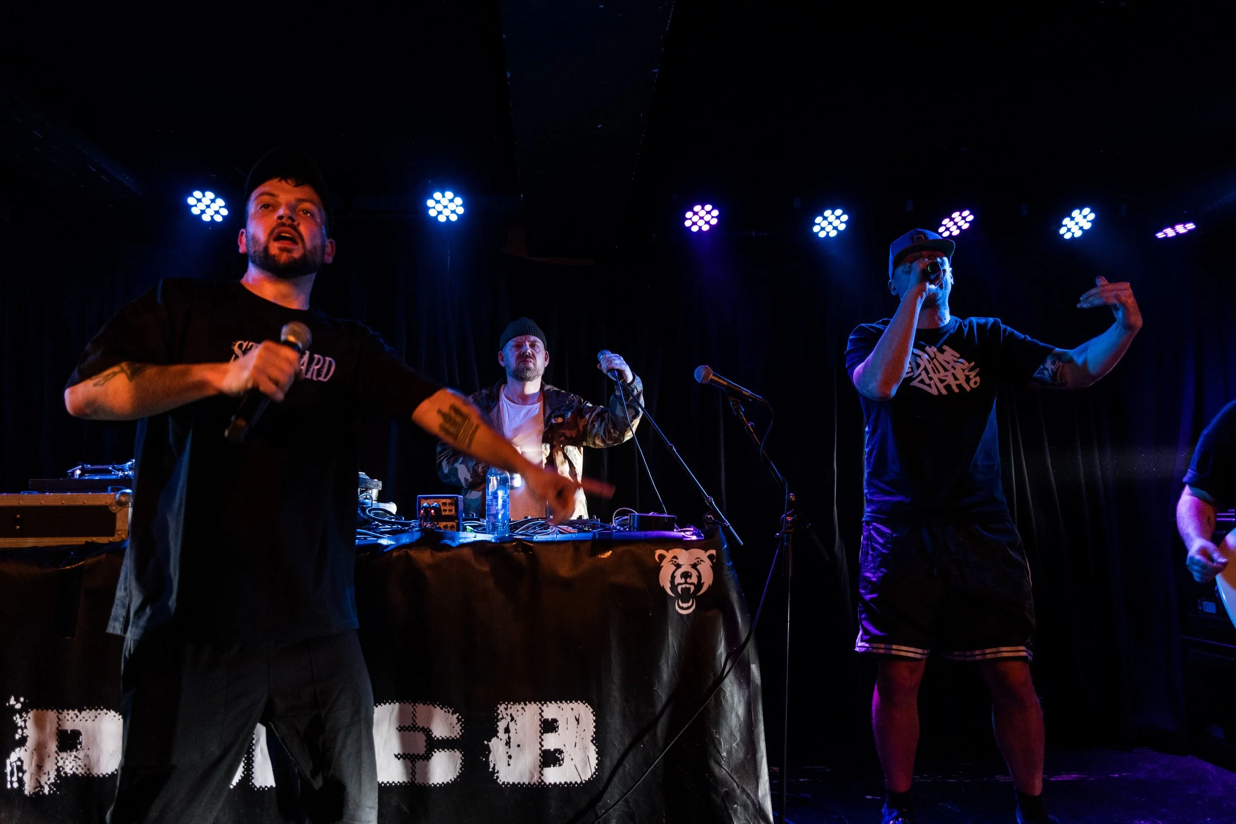 Three men performing on stage with music equipment, one holding a microphone, with blue stage lights and a black curtain backdrop. Bygningen, Frit Løb, Cillian Mxrphy