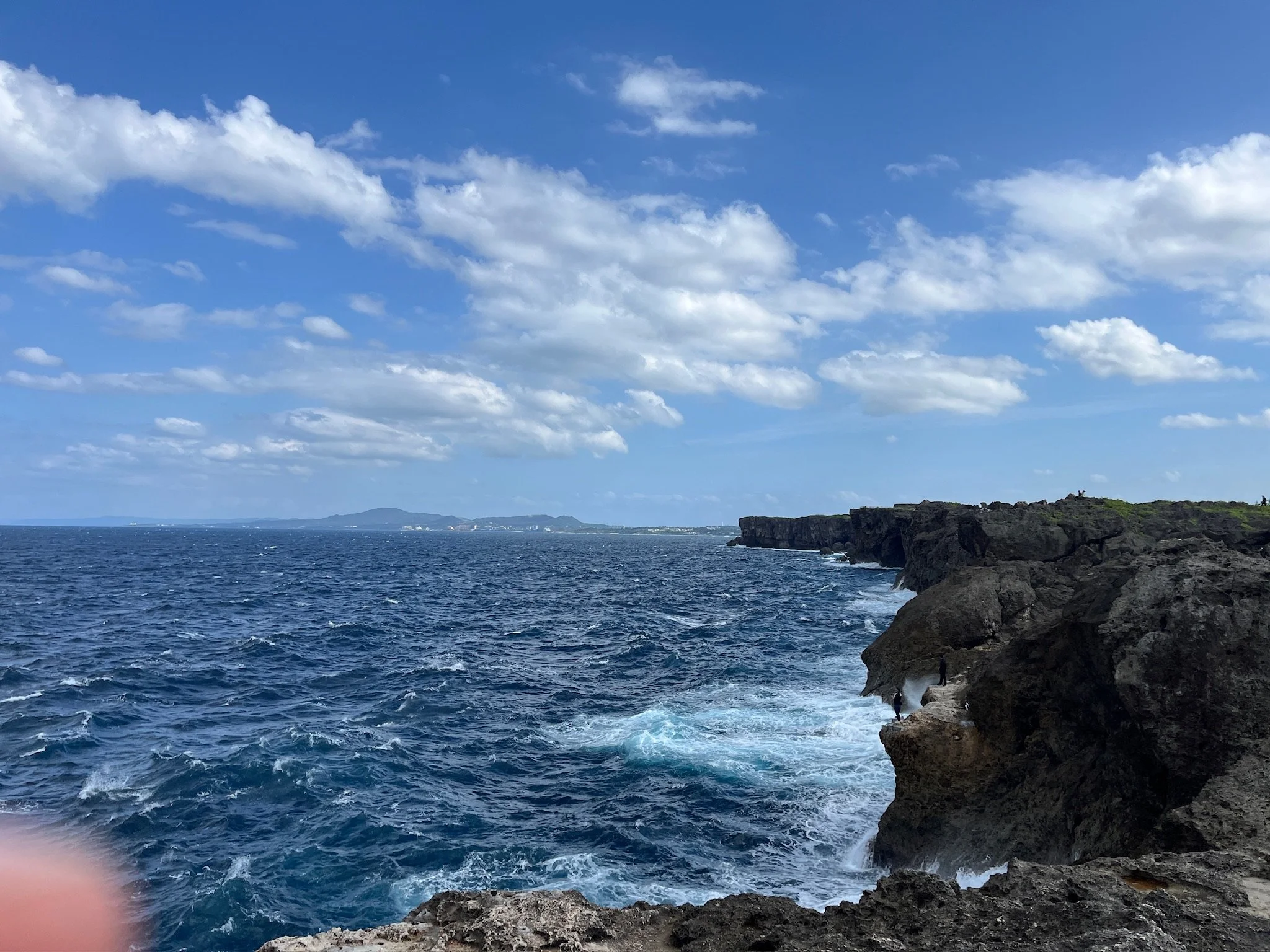 A scenic view of the ocean with rocky cliffs on the right side and a partly cloudy blue sky overhead in Okinawa, Japan
