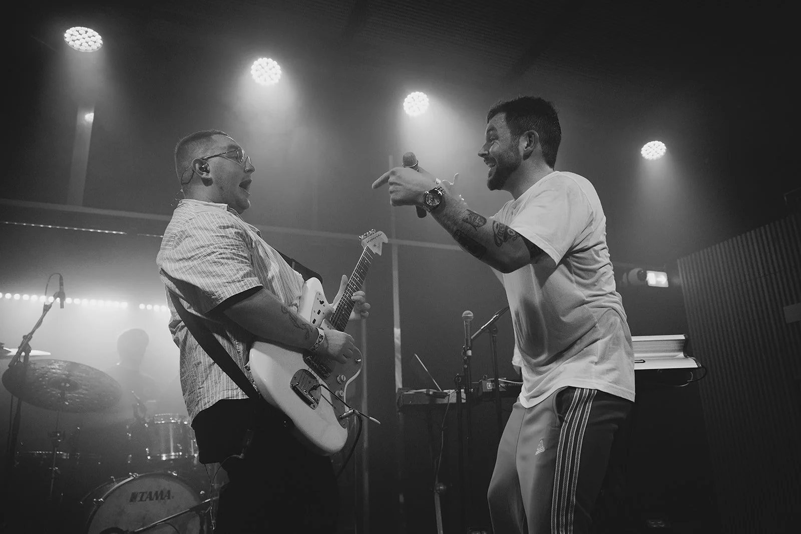Two musicians on stage, one playing an electric guitar and the other singing into a microphone, with band equipment in the background. Billede af Tinus Buus.