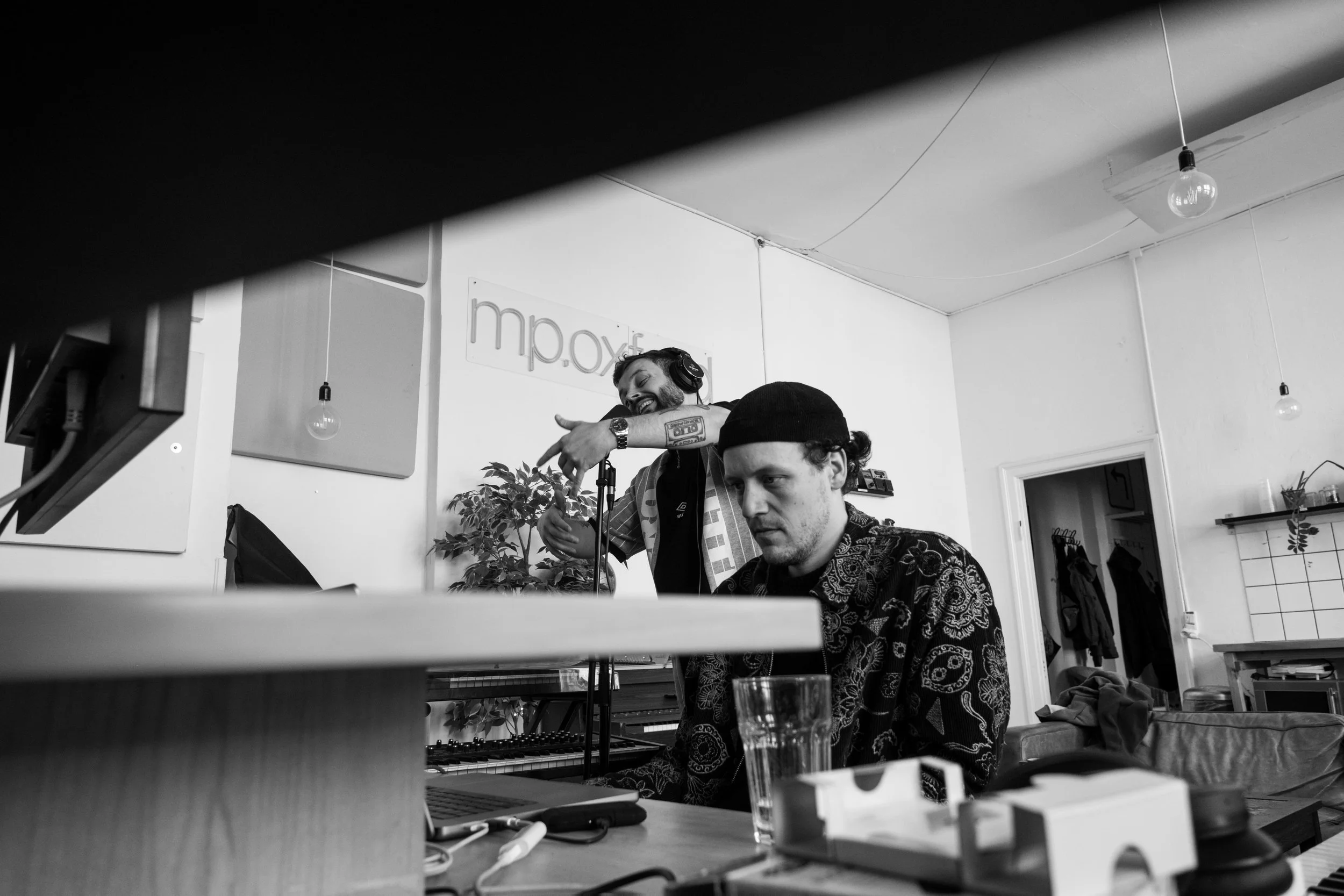 Two men in a recording studio, one wearing headphones and singing or speaking into a microphone, the other sitting at a keyboard, focused on the equipment, with various accessories and a glass of water on the table, in a minimalist room. Tinus Buus.