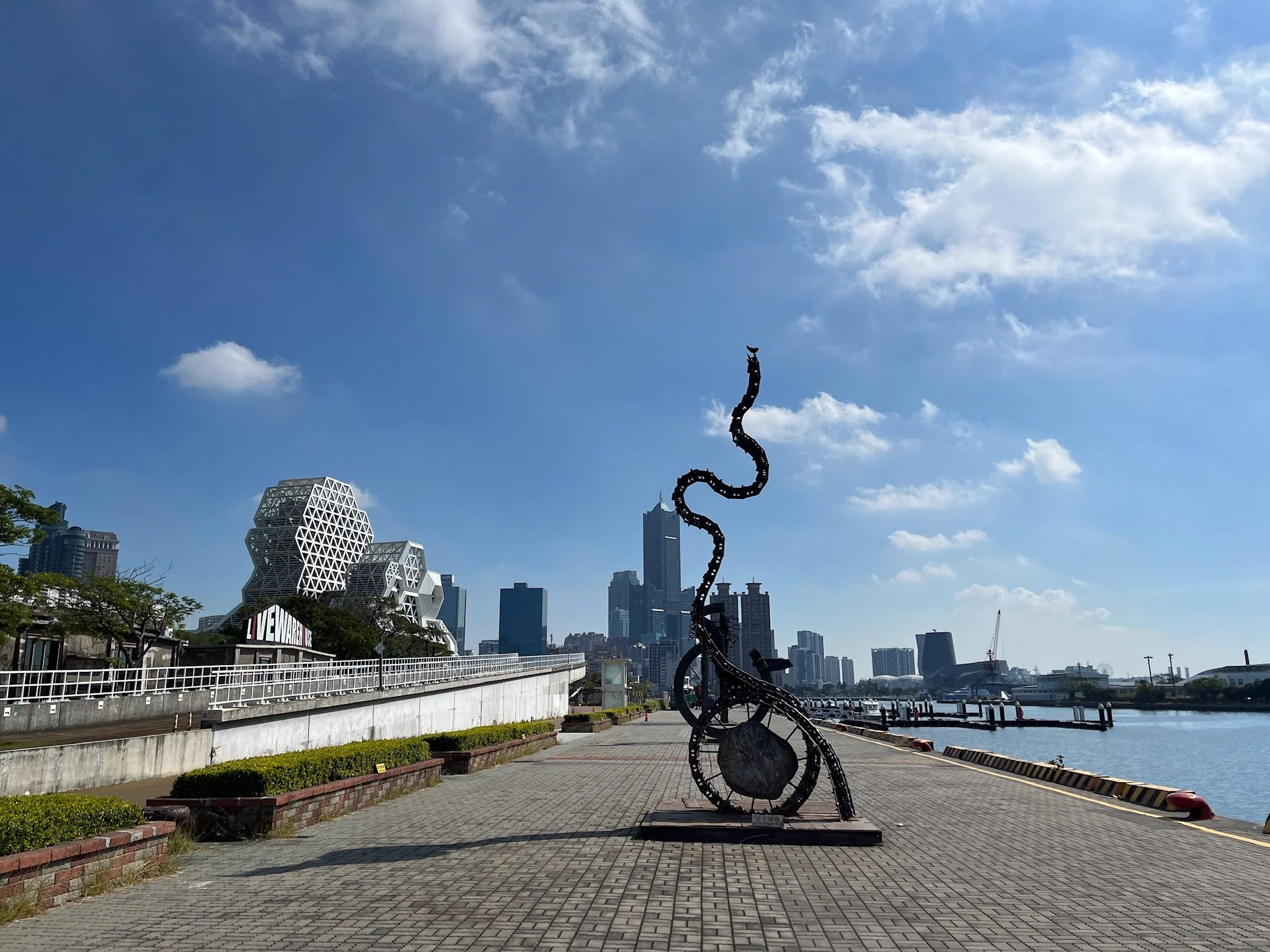 Sculpture of a musical note with a snake shape, located on a sidewalk along the South China Sea, with city buildings and a blue sky in the background of Kaohsiung, Taiwan.
