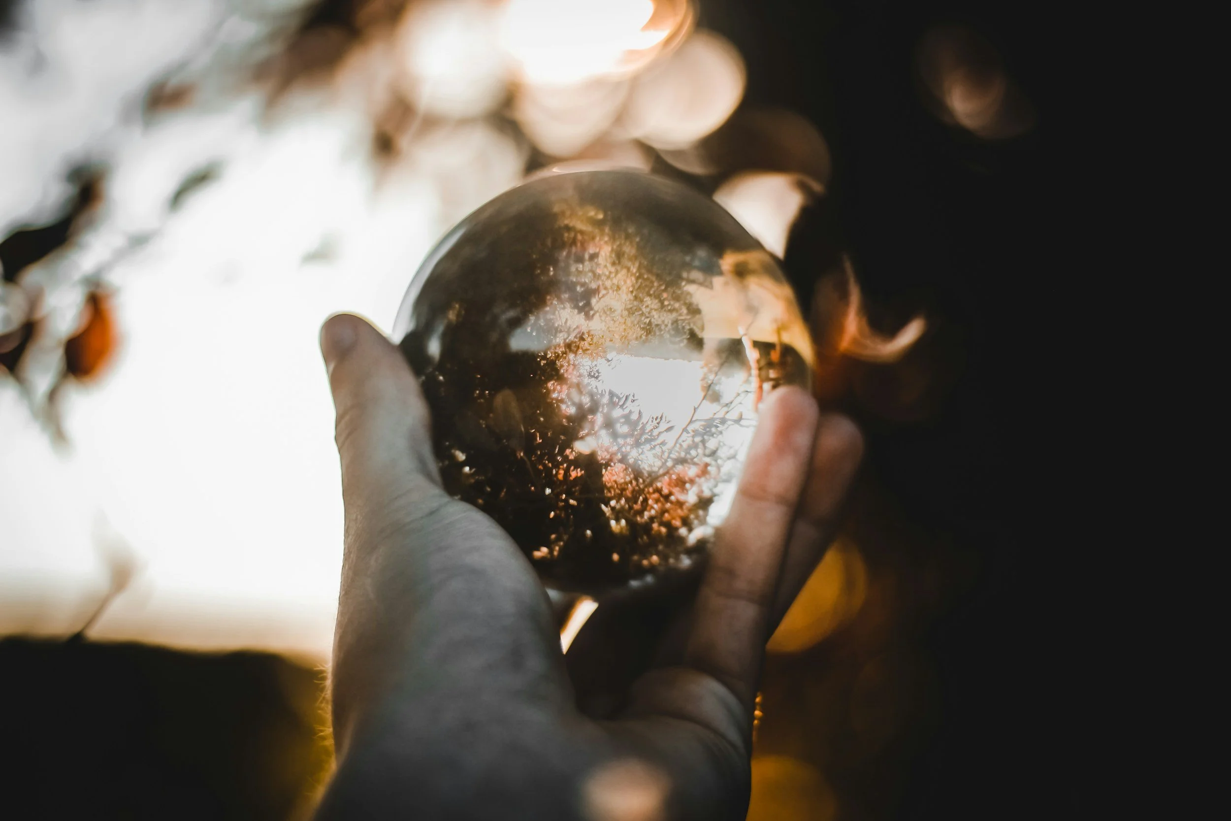 Main tenant une boule de cristal, reflet de la nature et du ciel à l'intérieur, avec un fond flou de lumière chaude.
