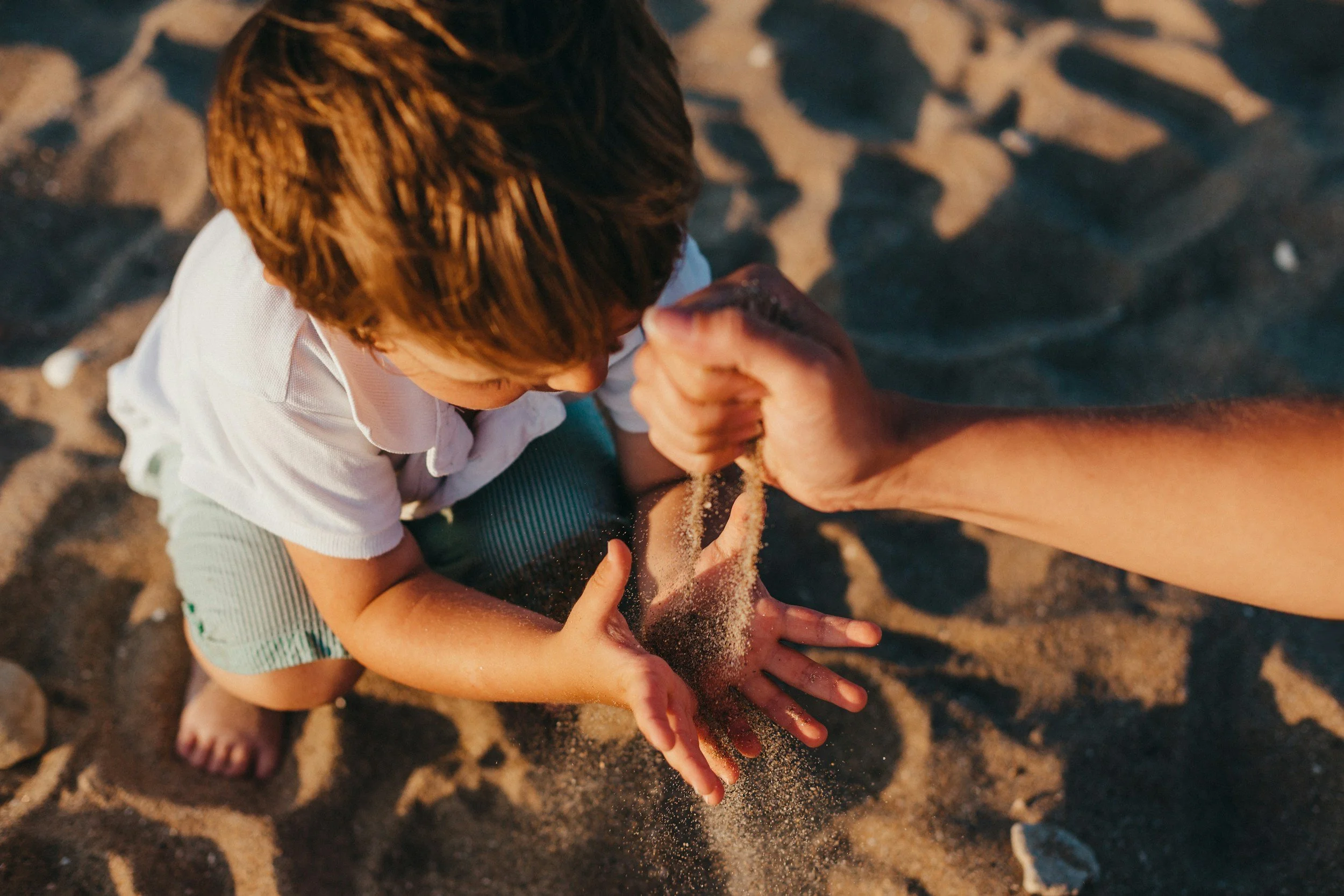 Un enfant assis dans le sable, en train de se faire aider à recevoir du sable d'une main adulte, sur une plage, avec le soleil couchant