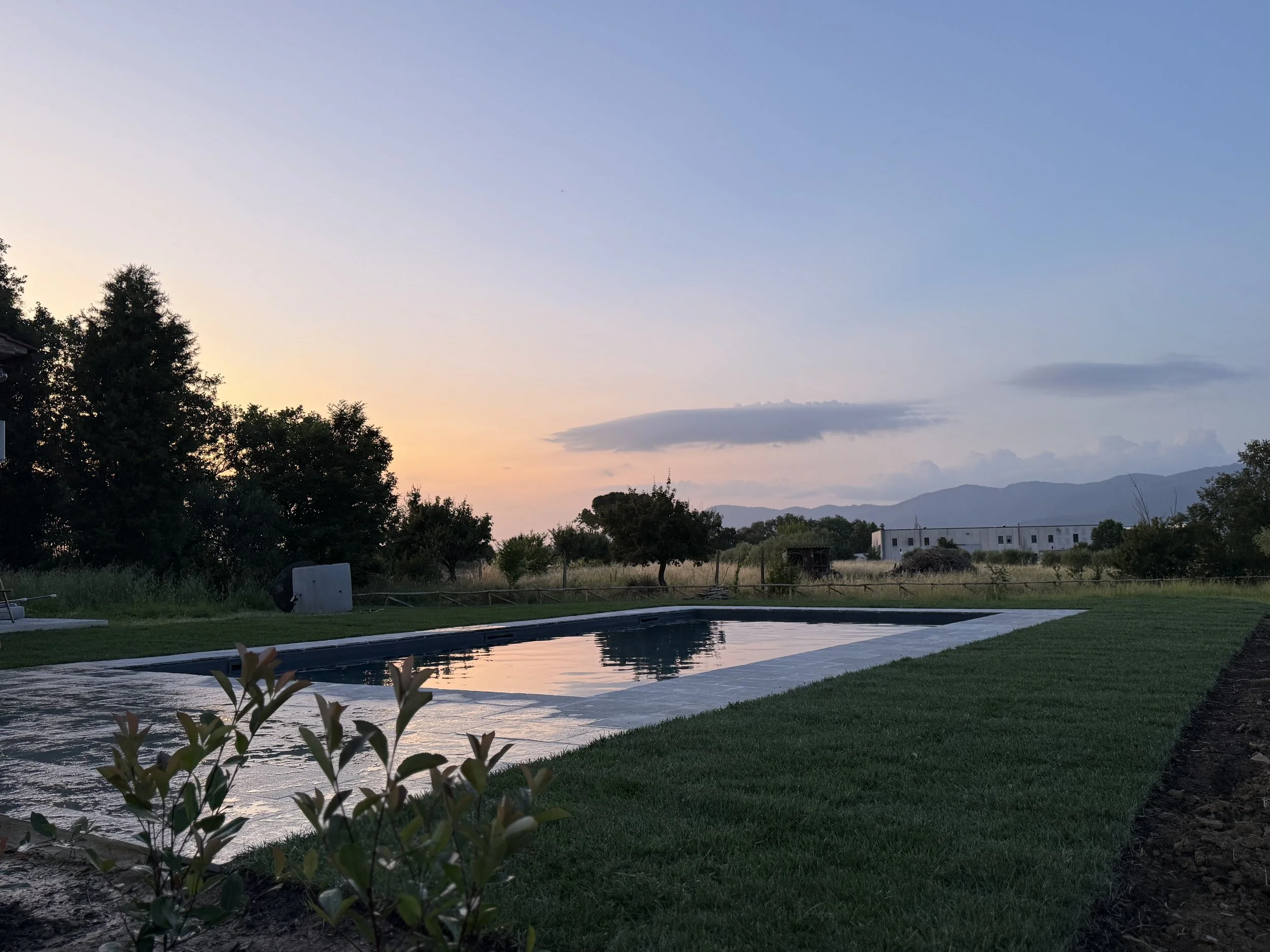 Swimming pool surrounded by green grass and trees at sunset with mountains in the background.