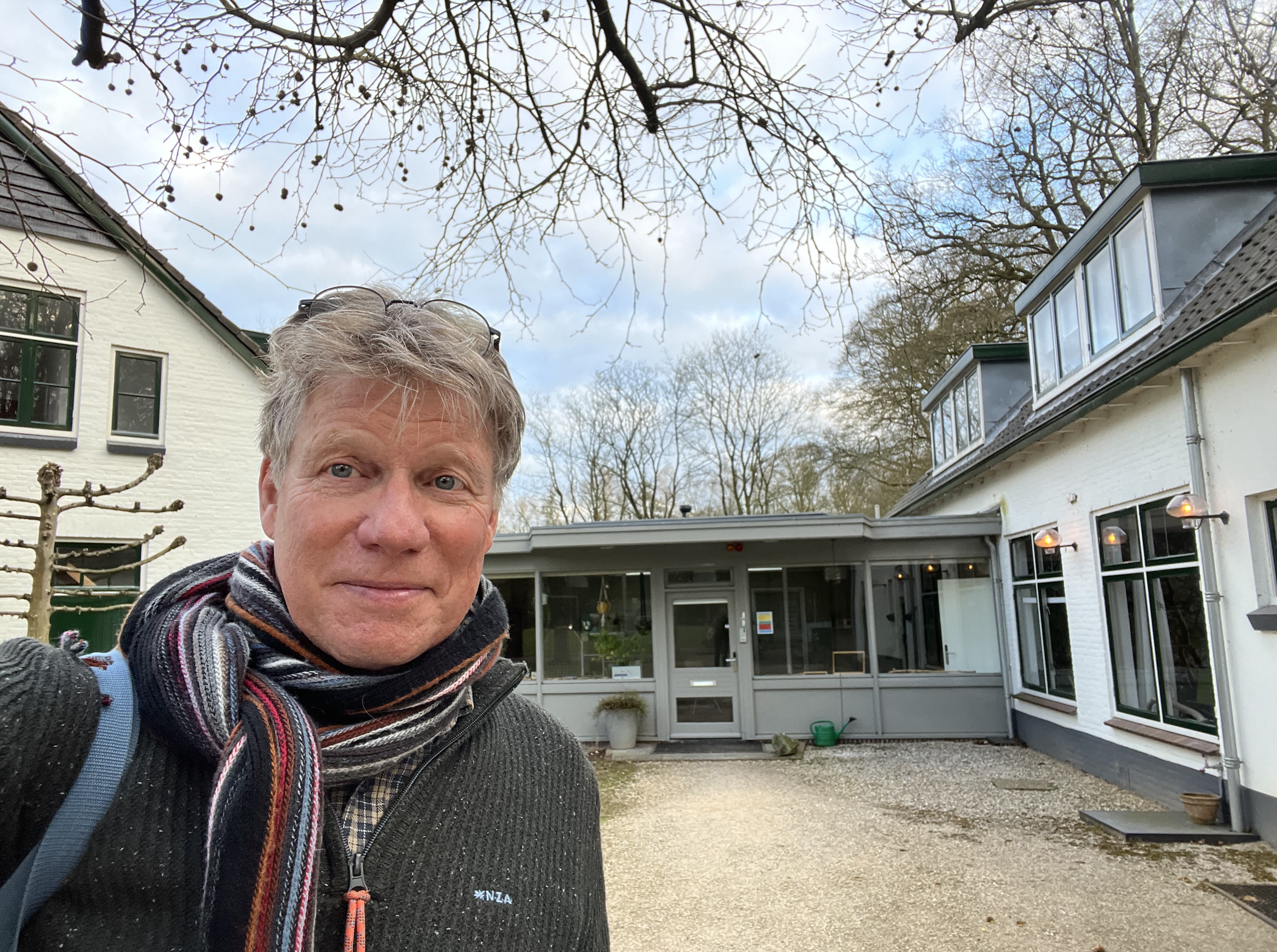 Een man neemt een selfie voor een wit huis met grote ramen en een patio, omringd door bomen zonder bladeren, tijdens een bewolkte dag.