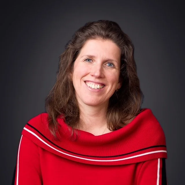 Portret van een vrouw met bruin haar, lachend, gekleed in een rode trui met witte details, tegen een grijze achtergrond.