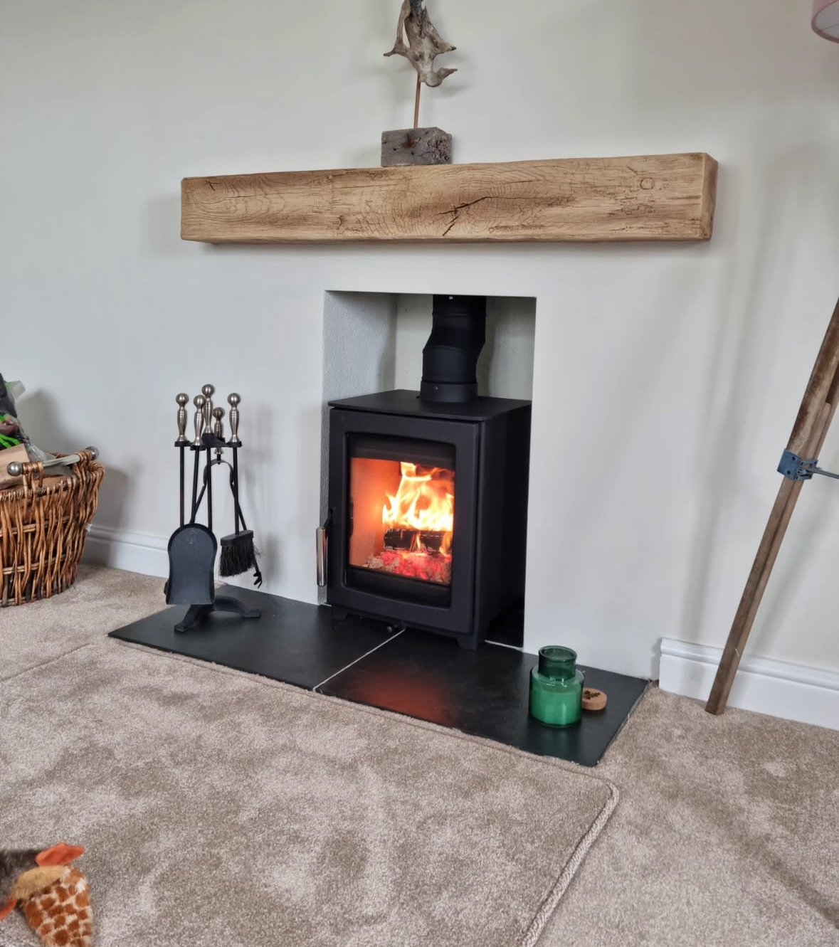 A wood-burning stove with a visible fire inside, set into a wall with a large natural wood mantel above it. To the left are fireplace tools, and to the right is a green candle jar on a black hearth pad on carpeted flooring.