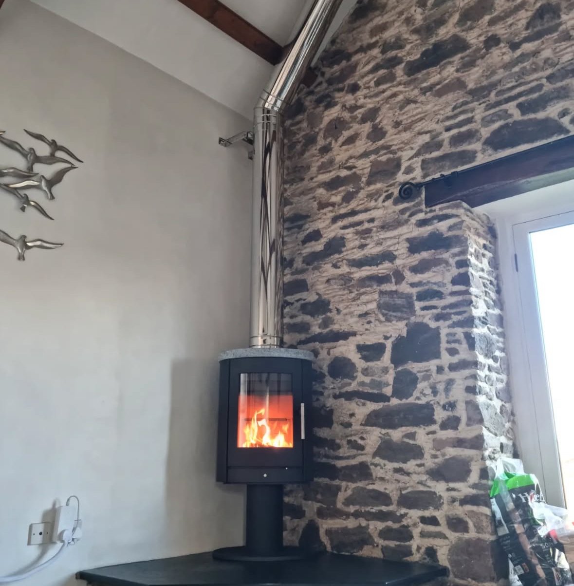 A small black stove with a glass door showing a fire inside, placed on a black surface, connected to a silver pipe chimney mounted on a textured stone wall, in a cozy interior with a window.