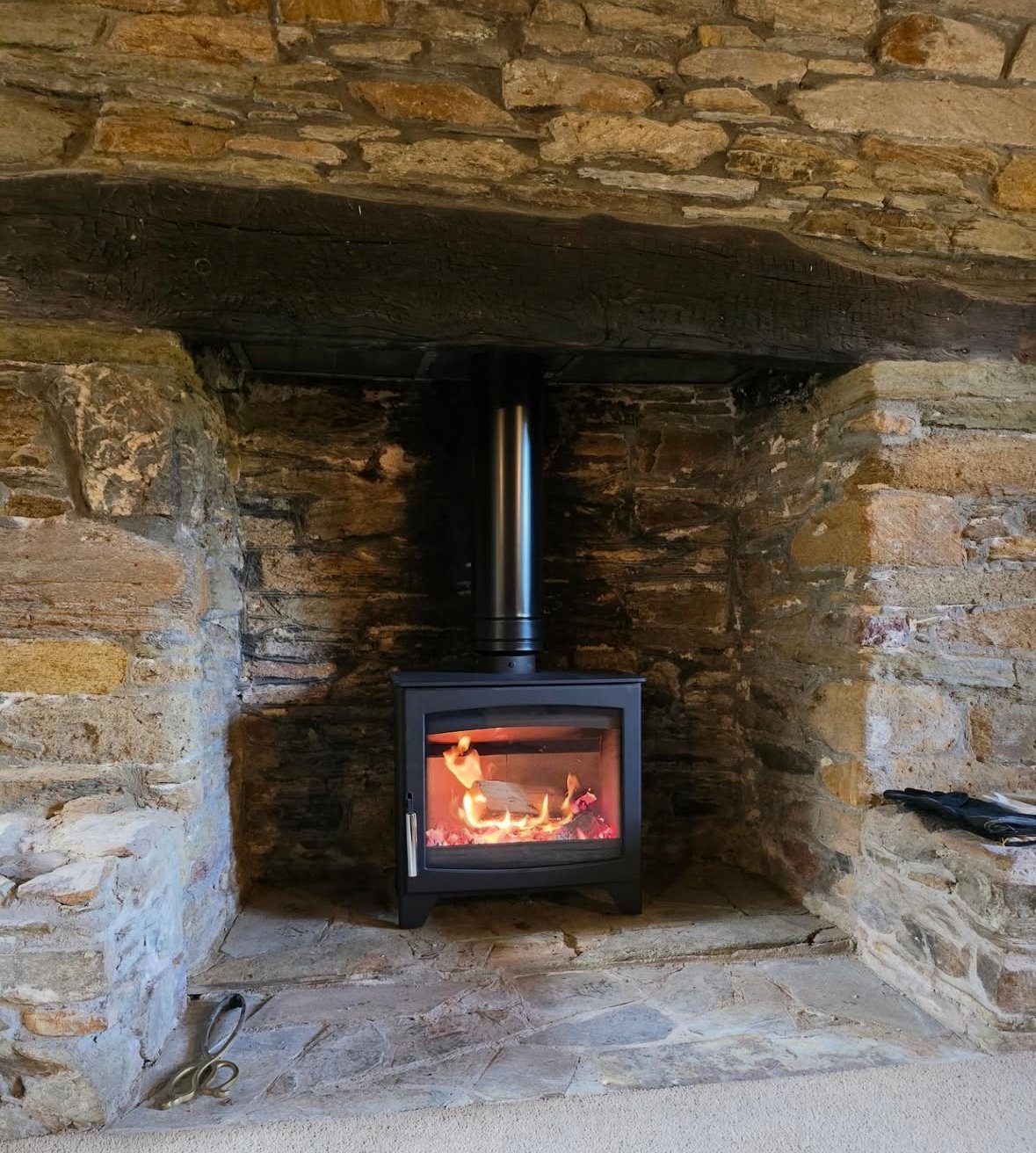 A wood-burning stove with flames inside, set in a stone fireplace with a brick and stone wall background.