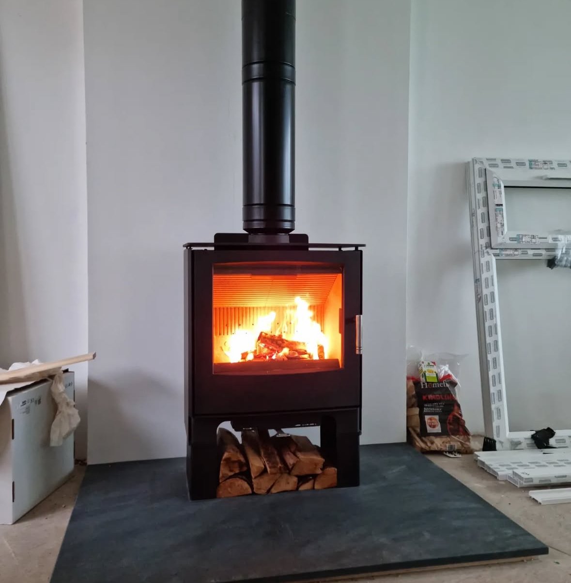 Wood-burning stove with a fire burning inside, with stacked logs underneath and construction materials nearby.