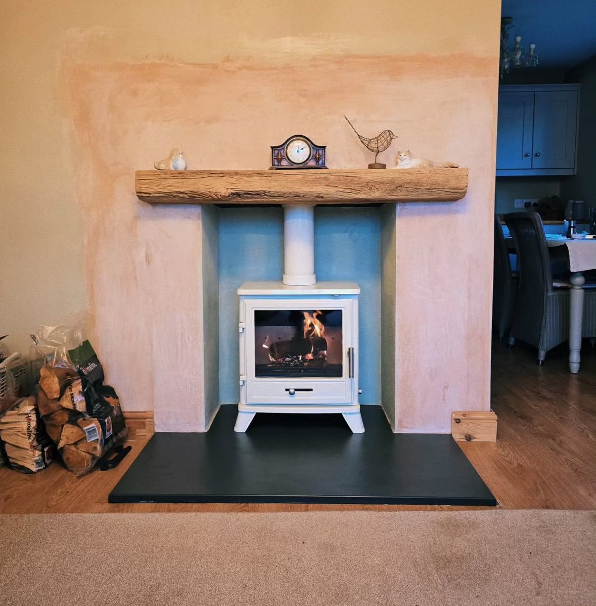 A cozy fireplace with a white stove, burning with flames, set against a textured pastel pink wall. A large rustic wooden mantel holds decorative items, including a clock and small sculptures. To the left, firewood is stacked in a basket. To the right
