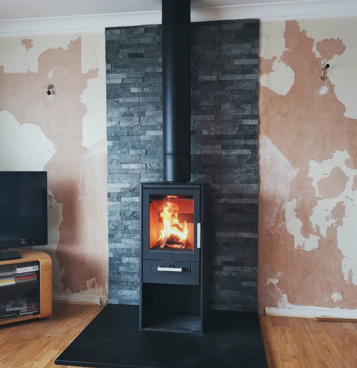 A modern wood stove with a visible fire, set against a wall with a black stone panel in the center, flanked by areas of peeling paint on beige walls.