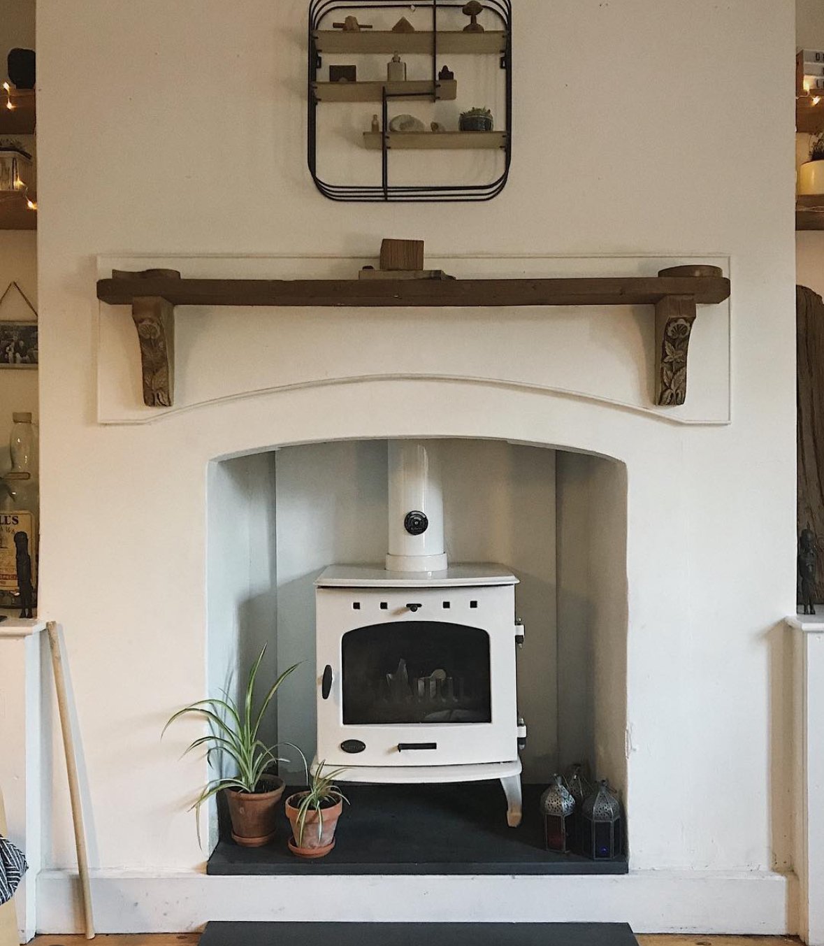 White fireplace with a vintage wood stove, two potted plants, and a wooden mantel shelf on a wall with a decorative black metal shelf above.