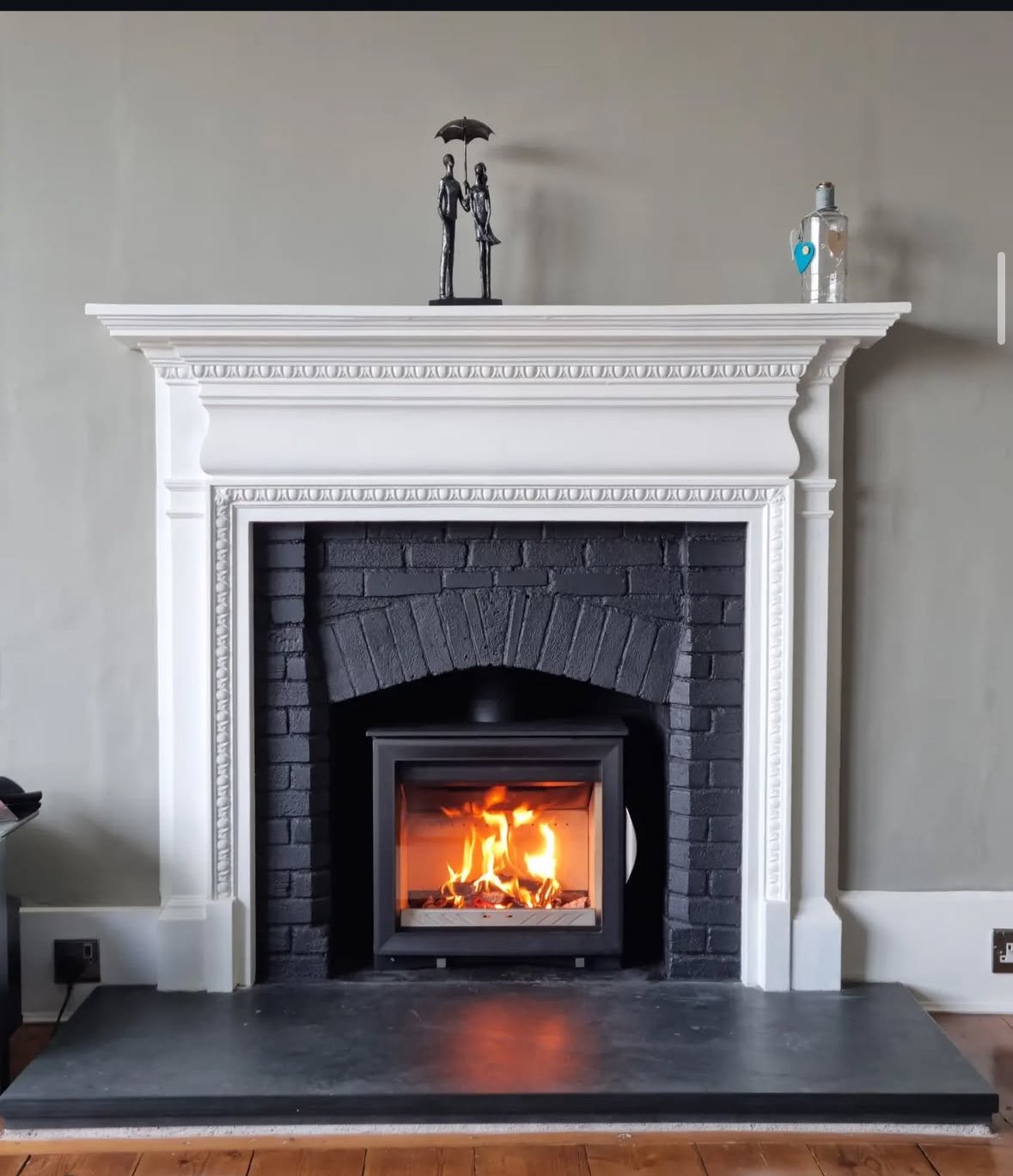White fireplace mantel with decorative molding, black brick interior, and a burning fire inside. Decorative figures with an umbrella and a bottle on top of the mantel.